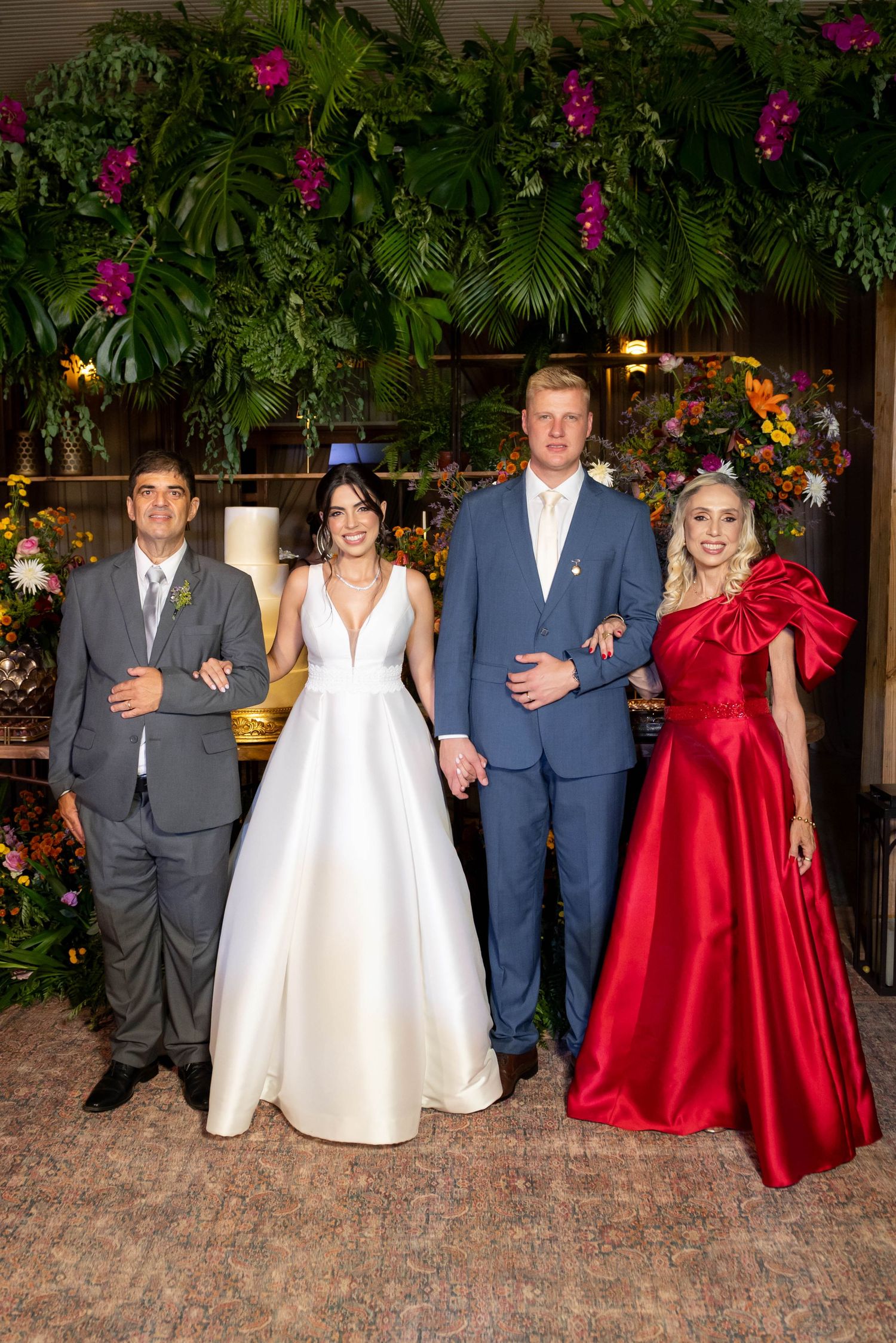 Wedding party of four posing elegantly, bride in white gown, groom in blue suit, guests in gray and red formal attire.