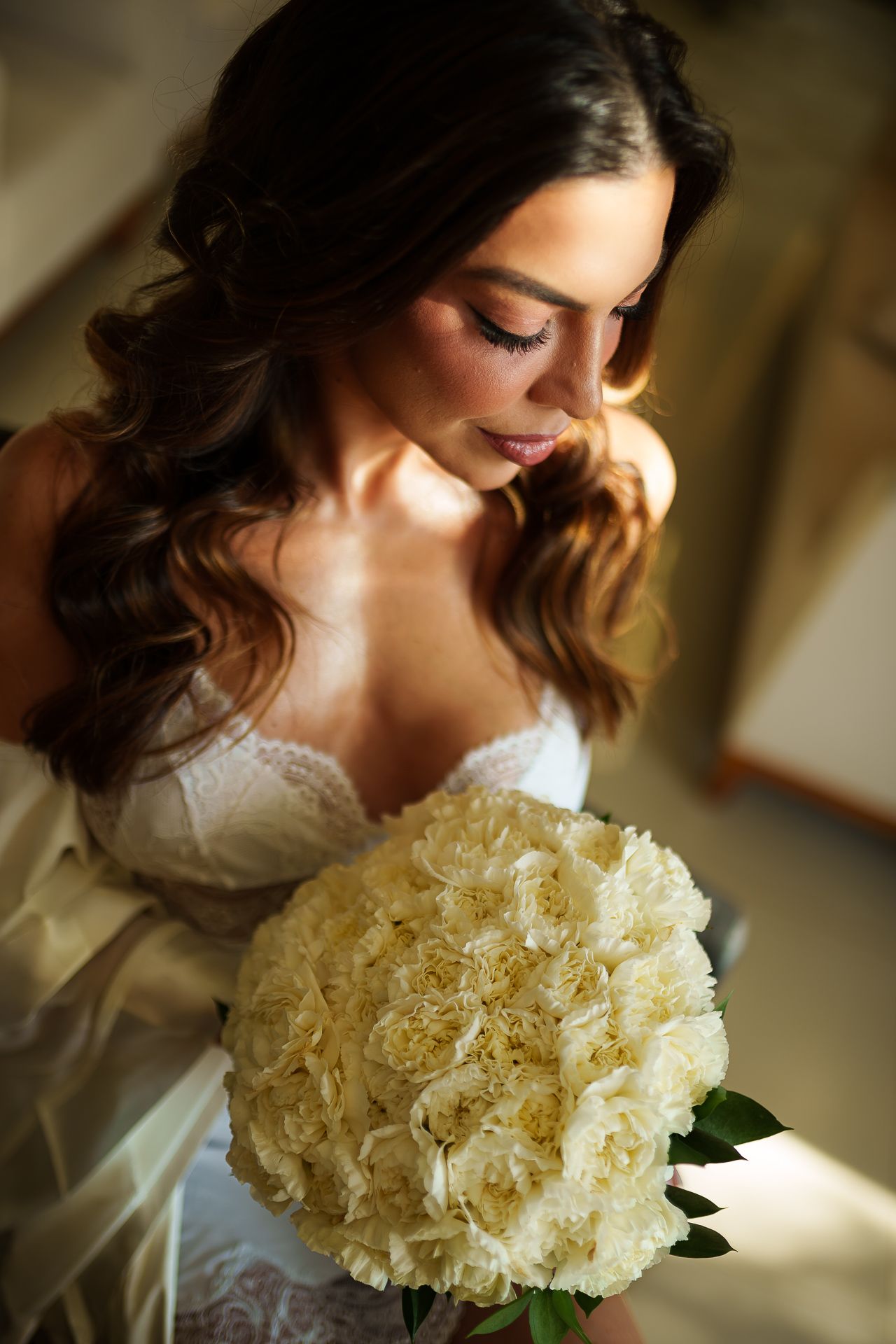 Bride in white lace lingerie holding large bouquet of cream roses, with soft natural lighting and elegant wavy hair.