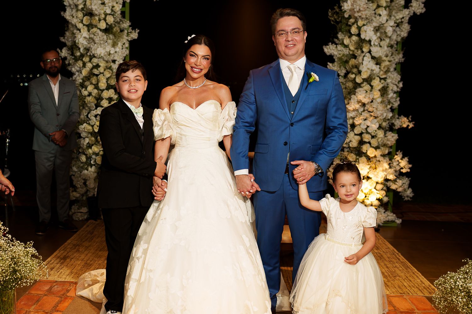 A bride in white gown and groom in blue suit pose with two children at an elegant floral wedding ceremony.