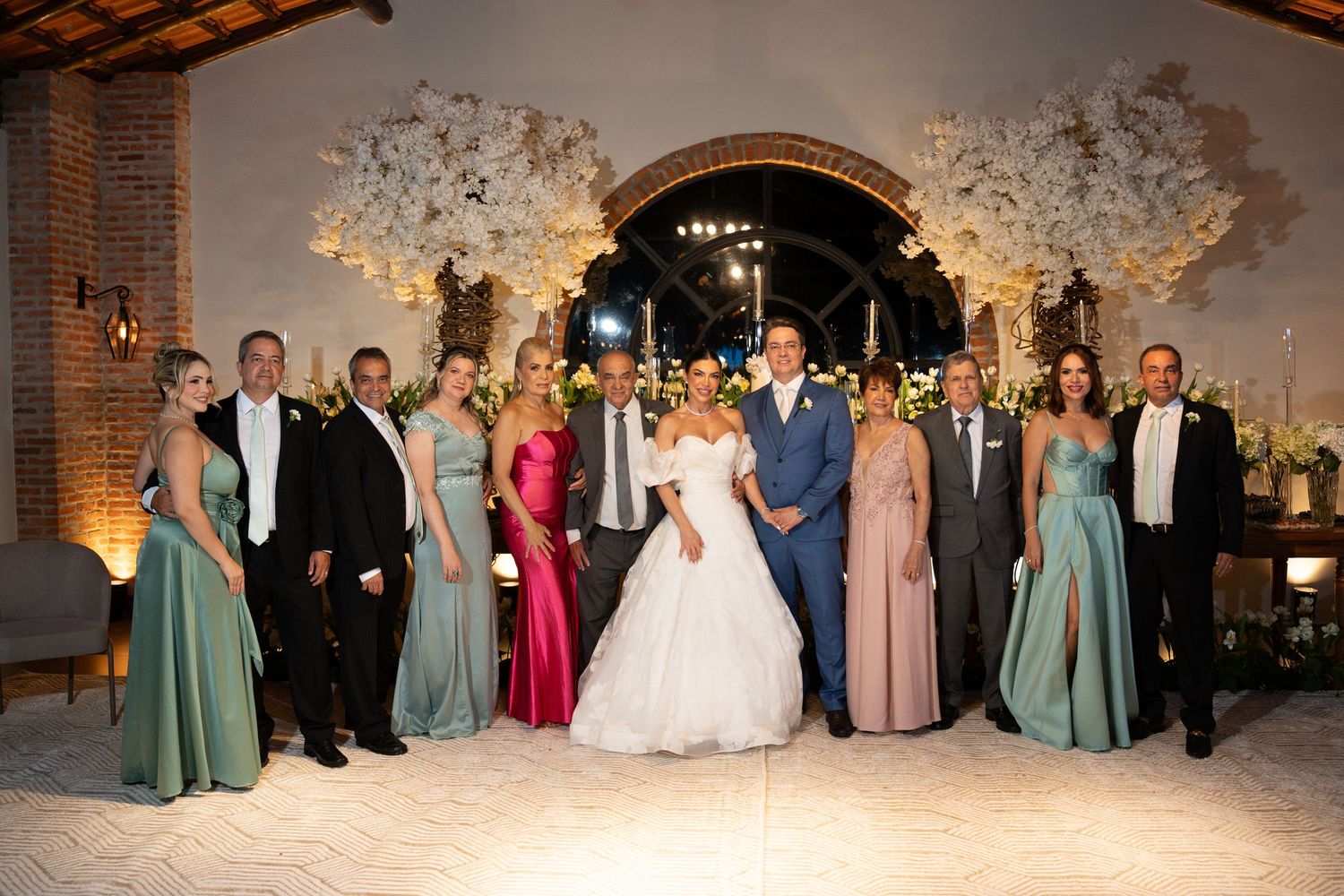 Wedding party poses in elegant venue with floral arch, bride in white gown, groom in blue suit, attendants in sage green.