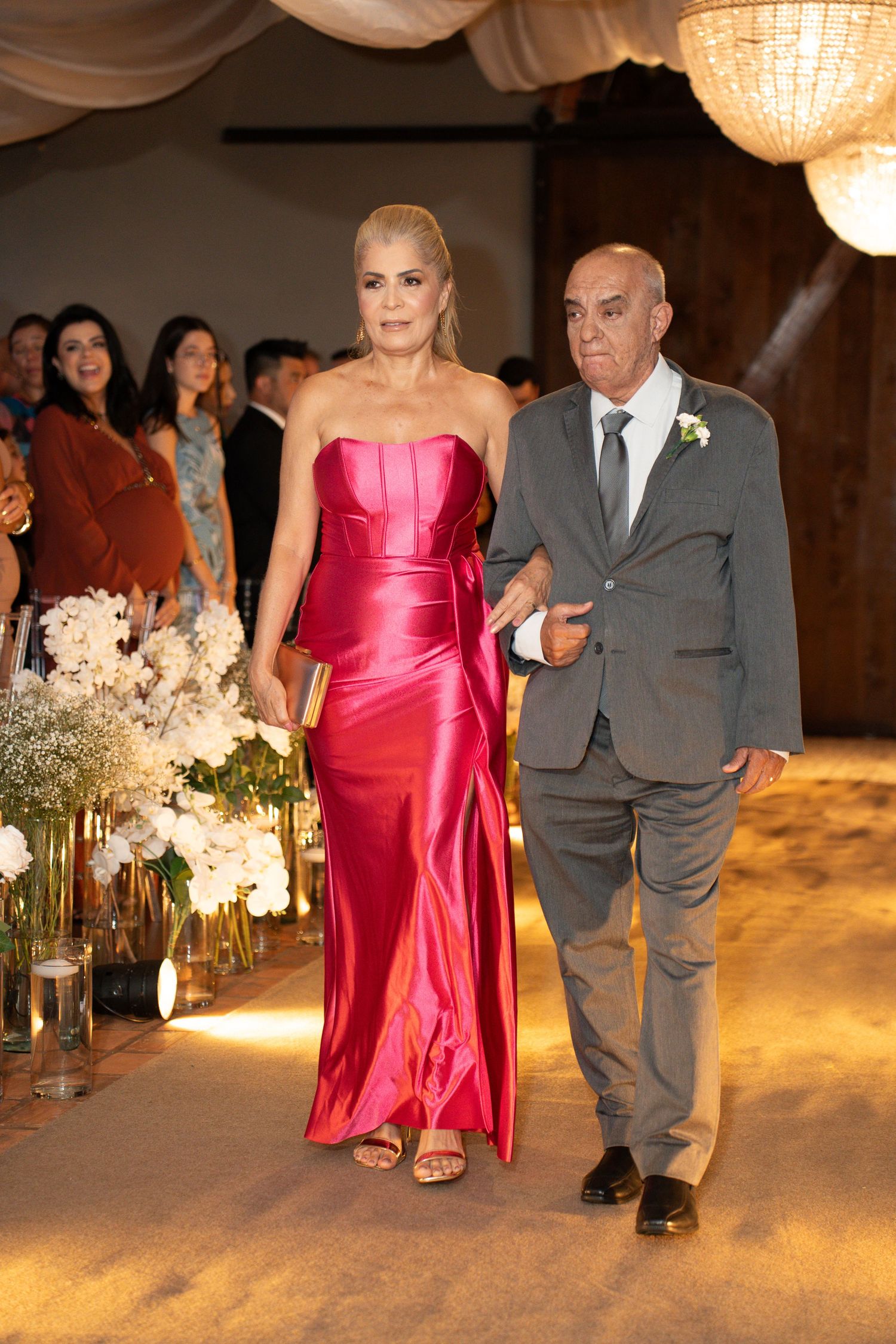 Elegant couple walking down wedding aisle, woman in strapless red satin gown, man in gray suit, floral arrangements lining the path.