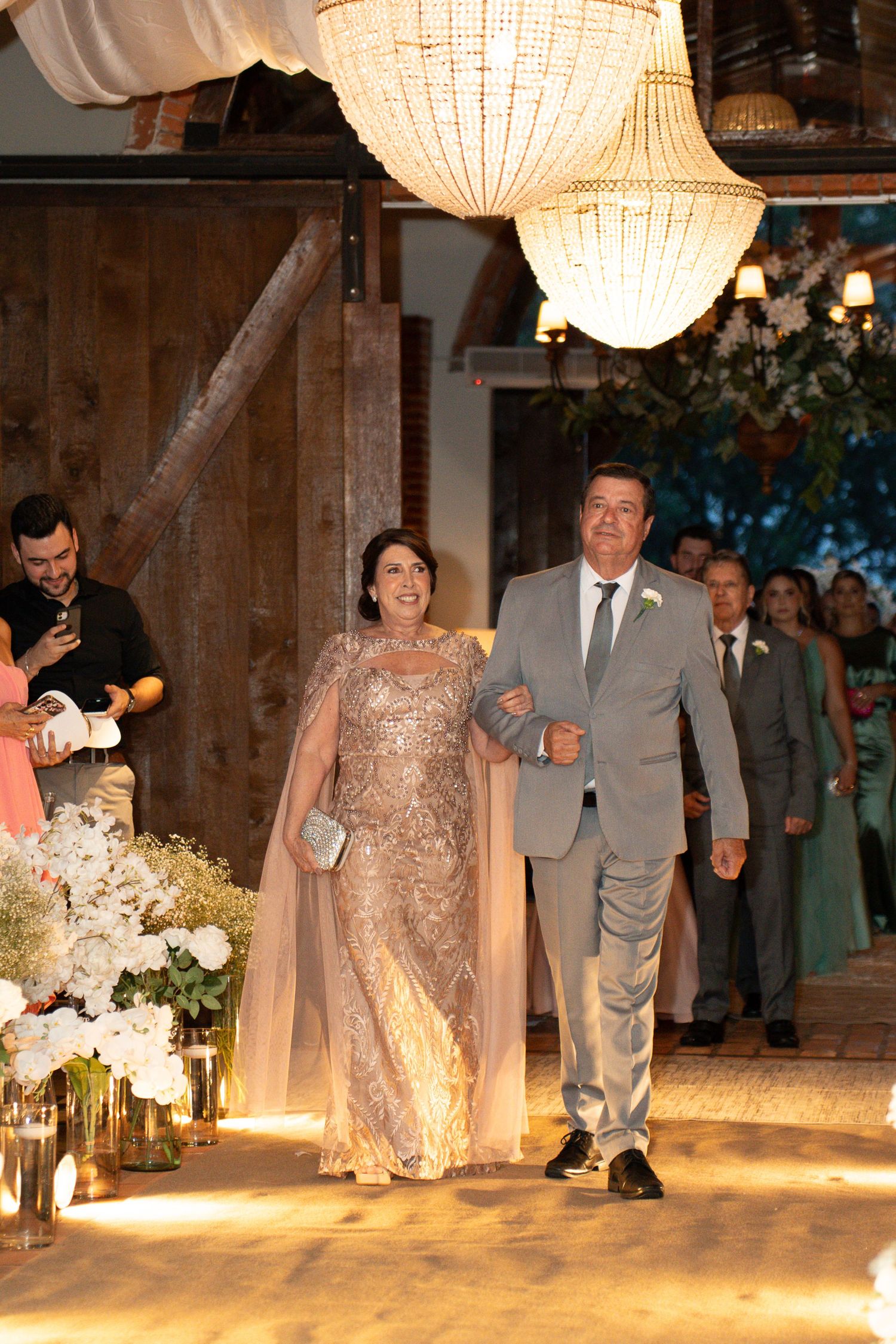 Elegant couple walking down wedding aisle, woman in gold gown with cape, man in gray suit, chandelier overhead.
