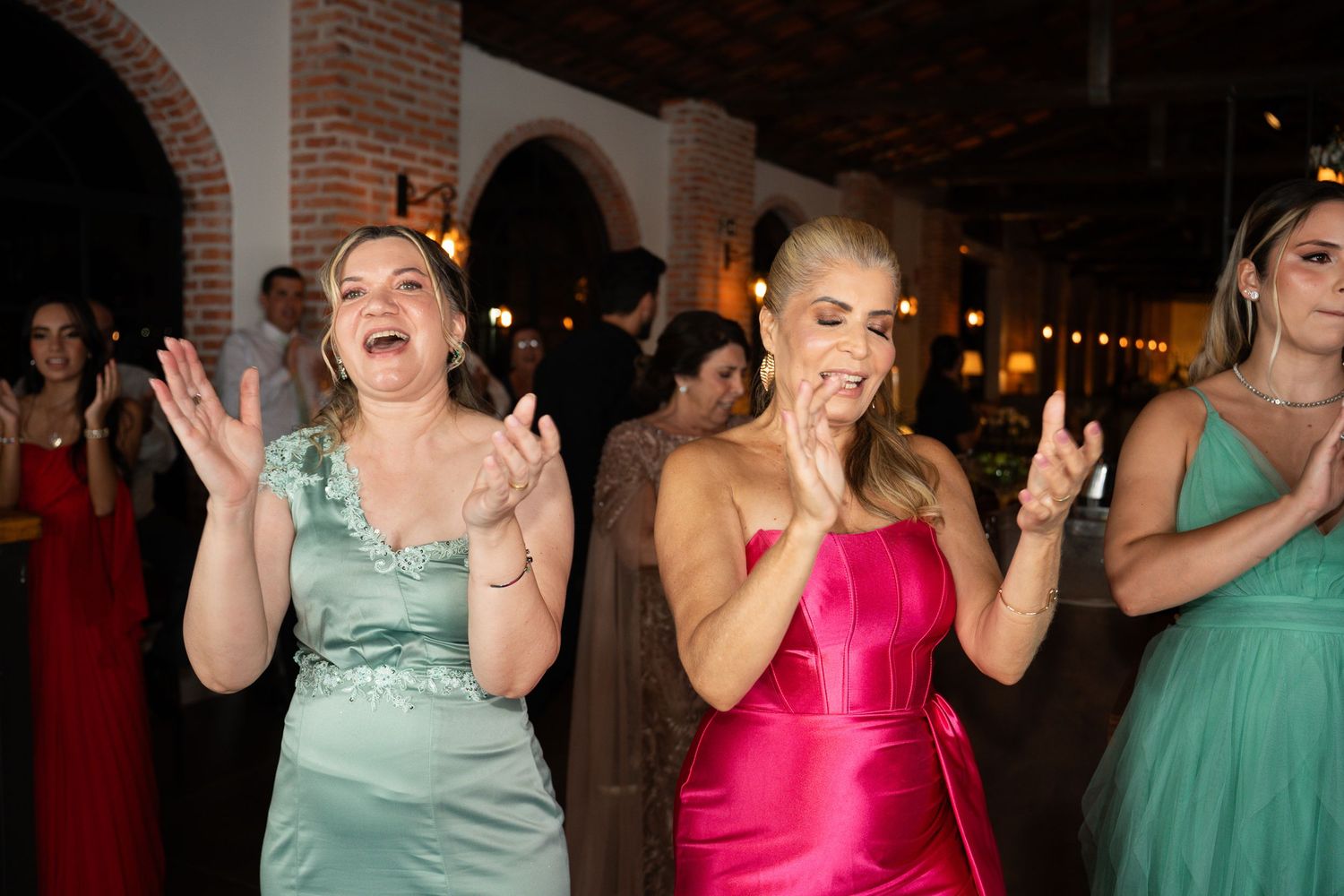 Women in colorful formal gowns clapping and dancing joyfully at an elegant indoor wedding reception.