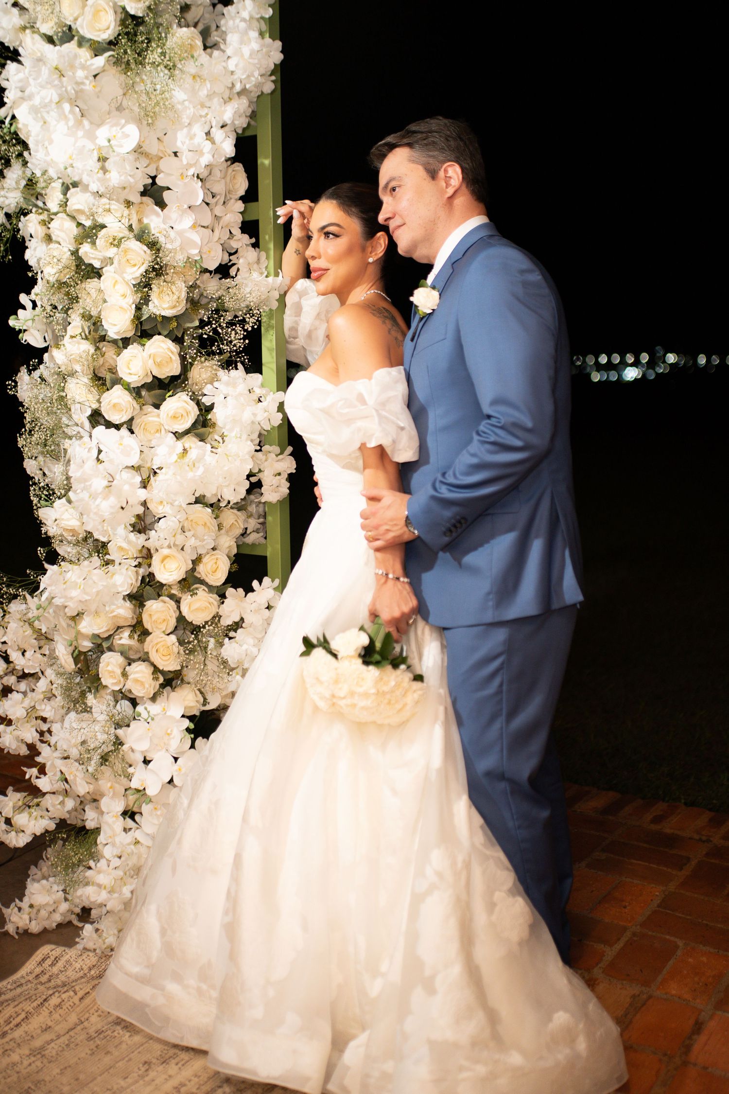 Bride in white off-shoulder gown and groom in blue suit pose by white floral arch at elegant evening wedding.