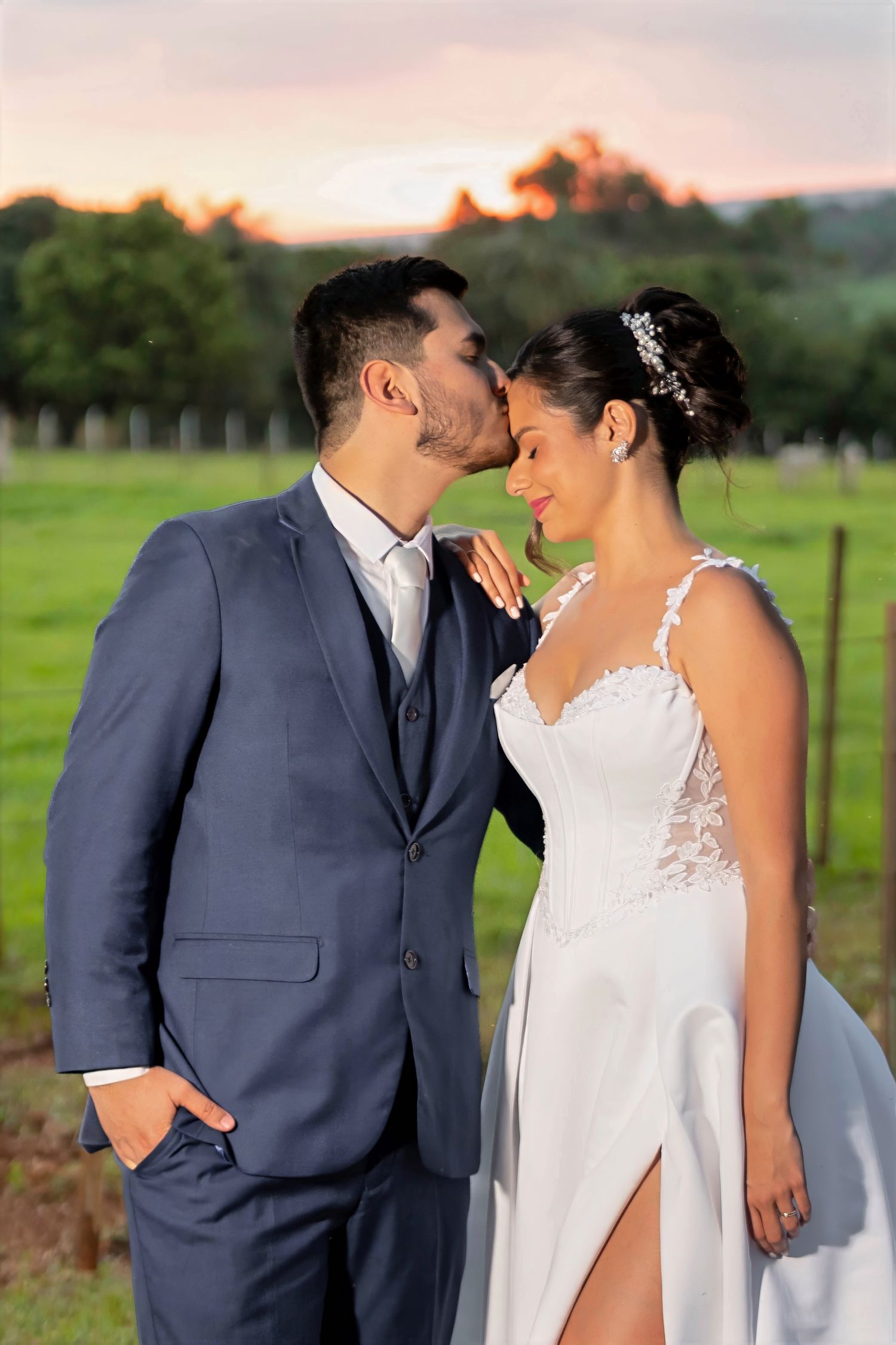 Groom in blue suit kissing bride in white lace wedding dress outdoors at sunset in green vineyard.
