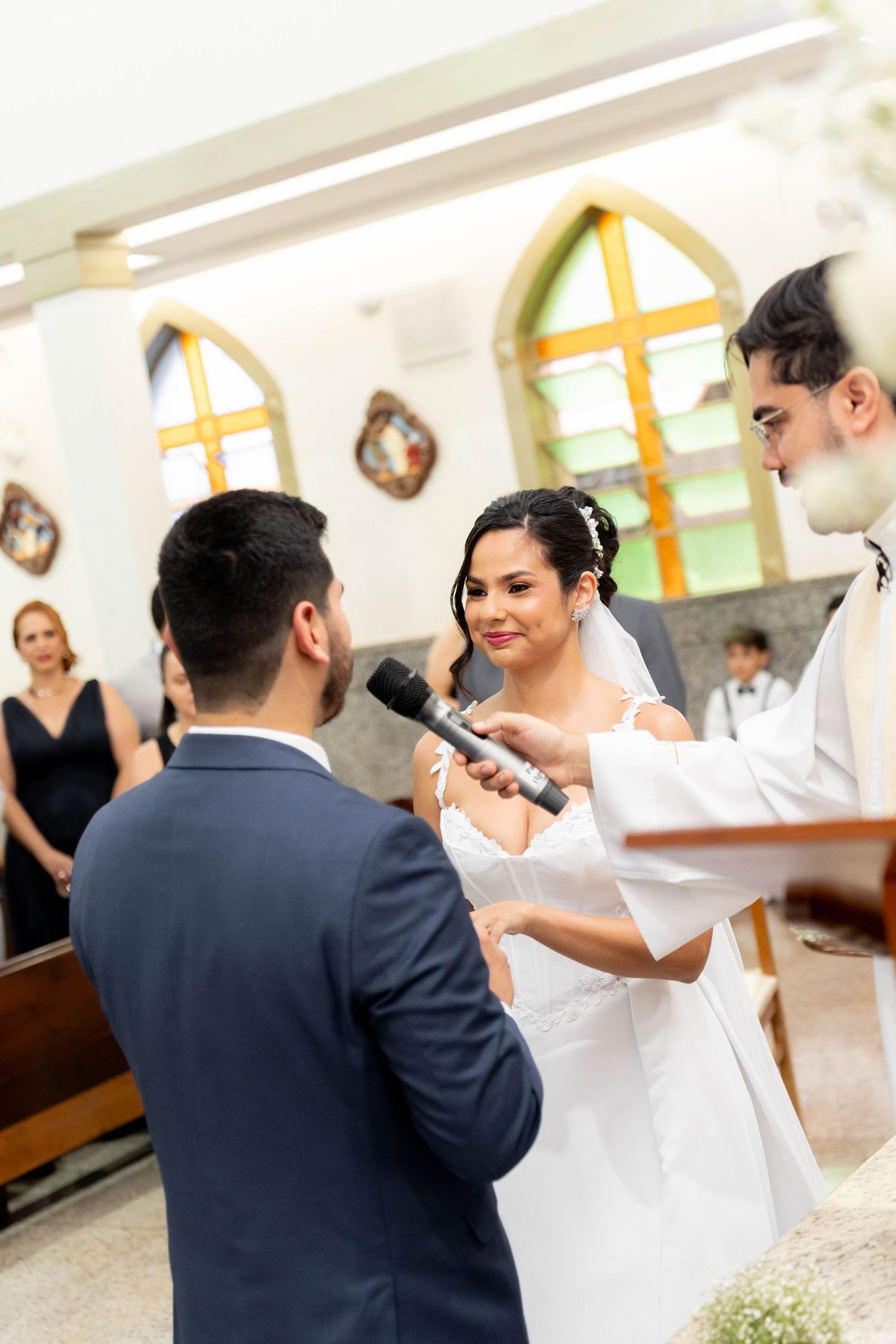 Bride and groom exchange vows at church altar during wedding ceremony, officiant holds microphone.
