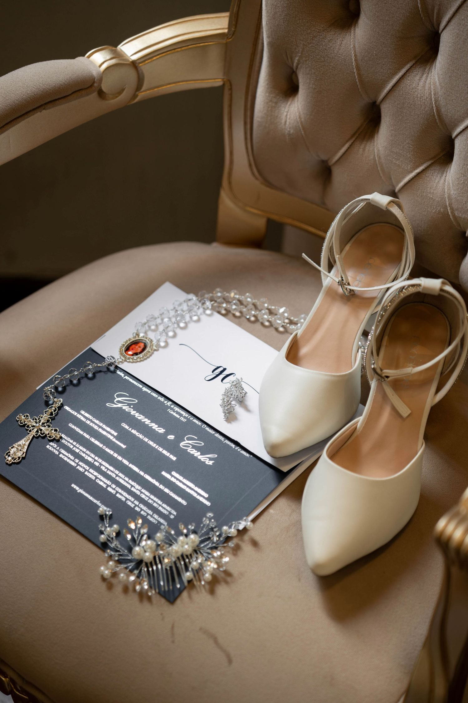 Elegant wedding flat lay with ivory heels, dark invitation suite, pearl necklace, and cross charm on a beige tufted chair.
