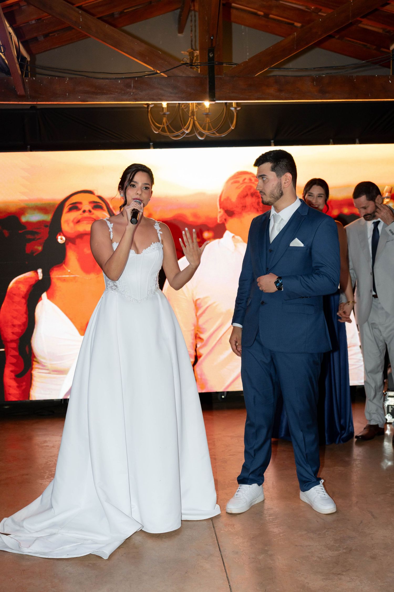 Bride in white gown speaks into microphone beside groom in navy suit at elegant wedding reception.
