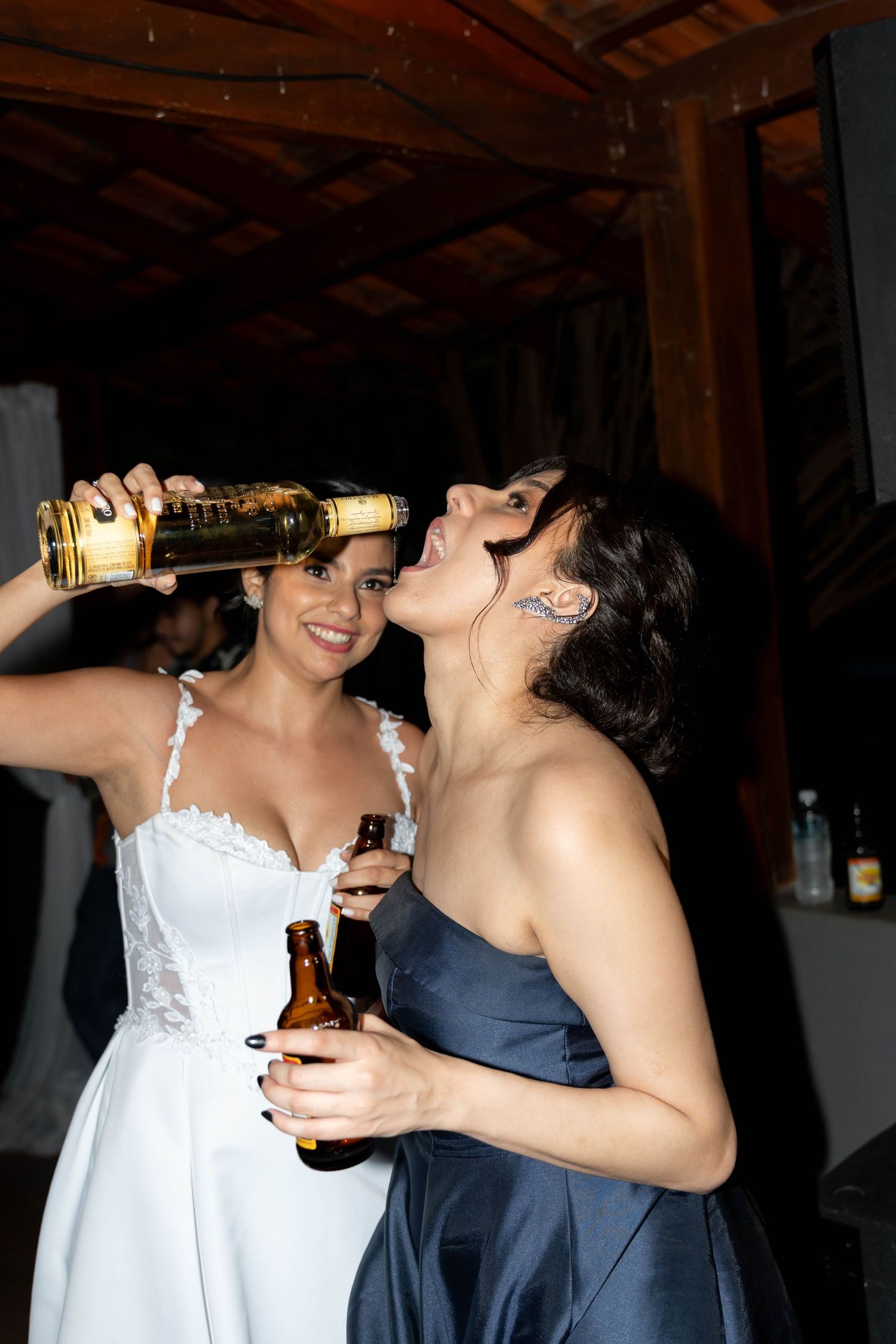 Bride in white dress and bridesmaid in blue gown drinking from bottles at a wedding reception celebration.