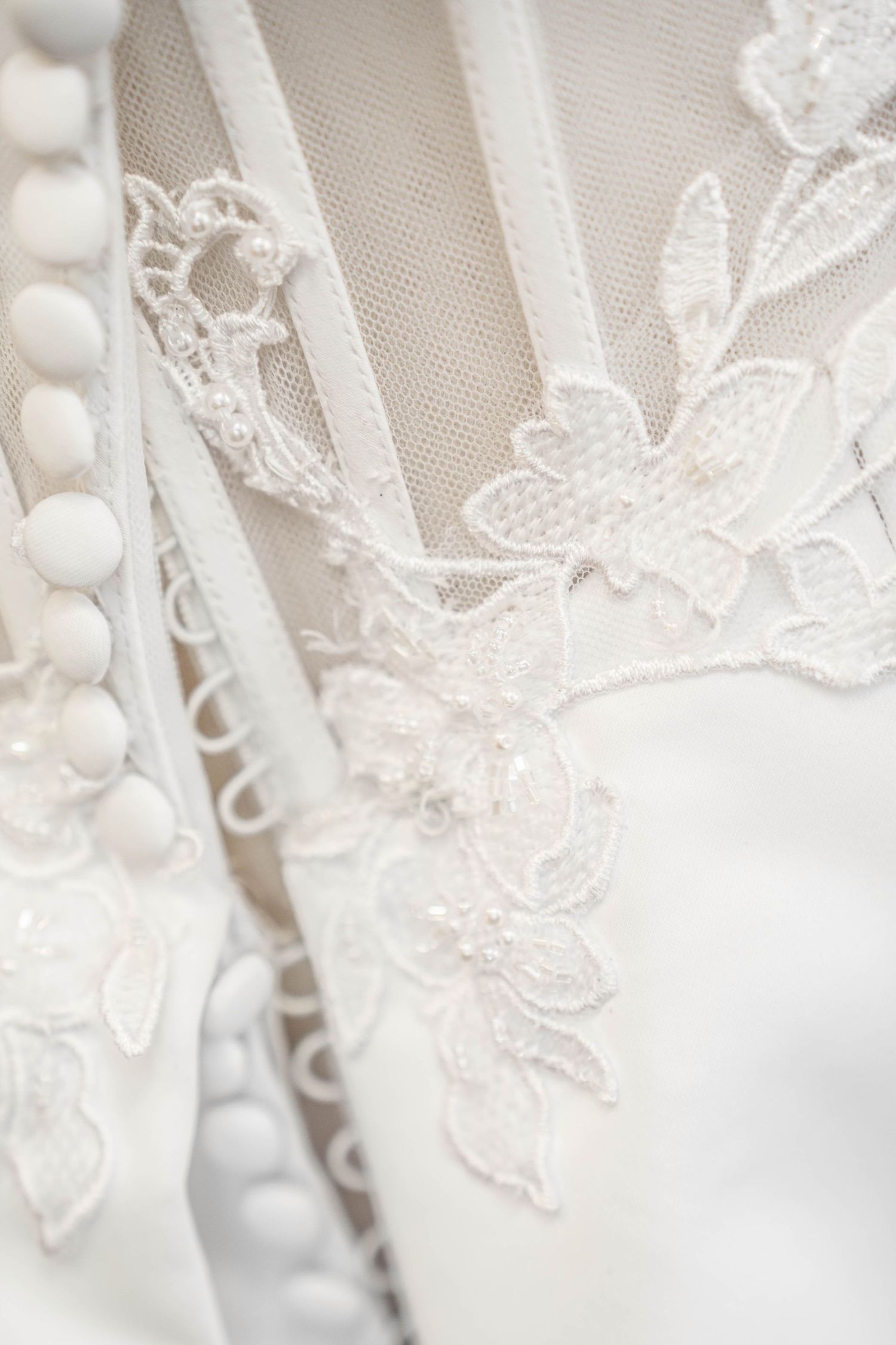 Close-up of elegant white wedding dress featuring intricate lace floral embroidery, sheer tulle panels, and delicate pearl button details.