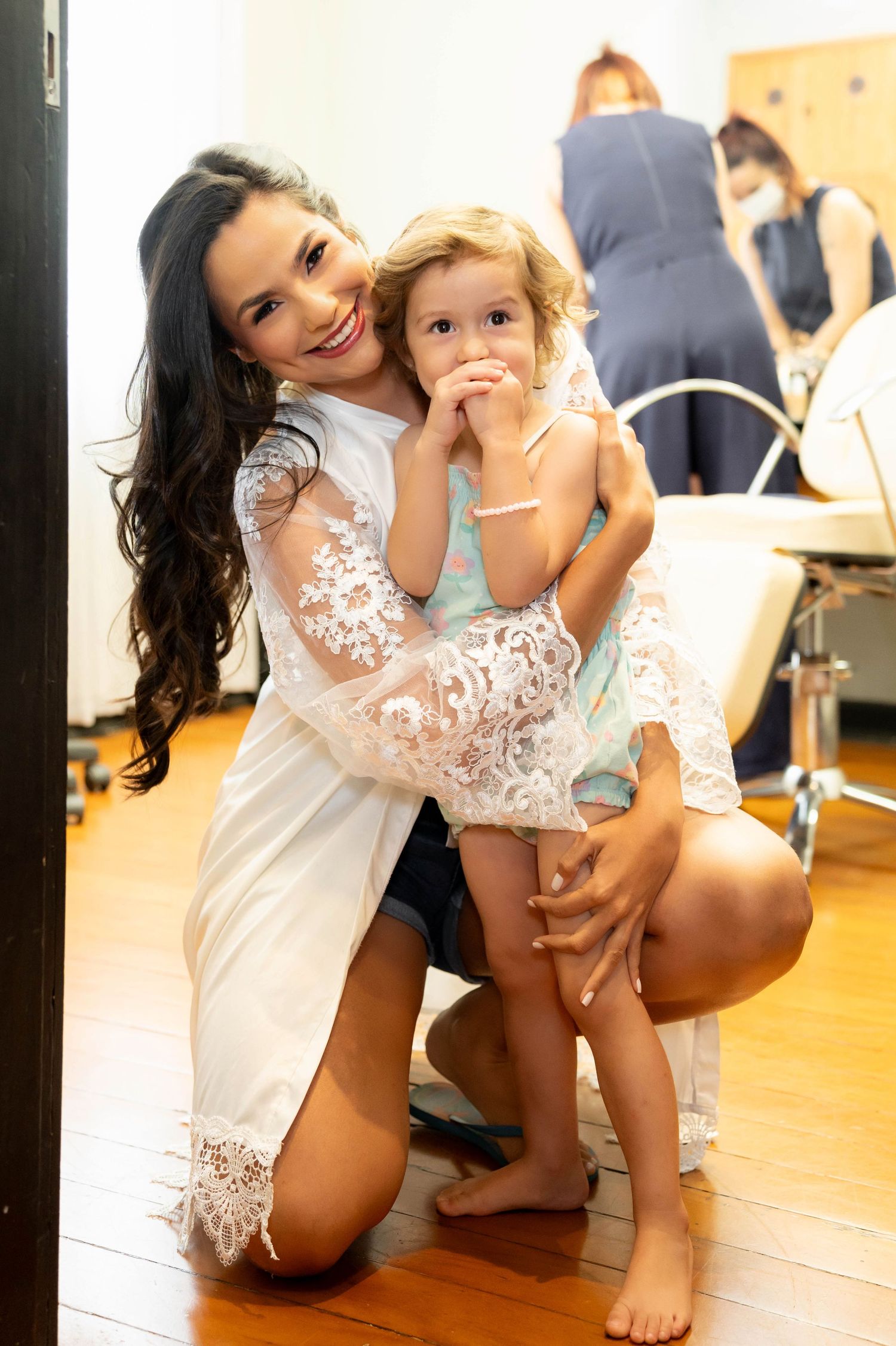 A smiling dark-haired woman in white lace robe kneels on wood floor hugging a curly-haired toddler girl in a salon.