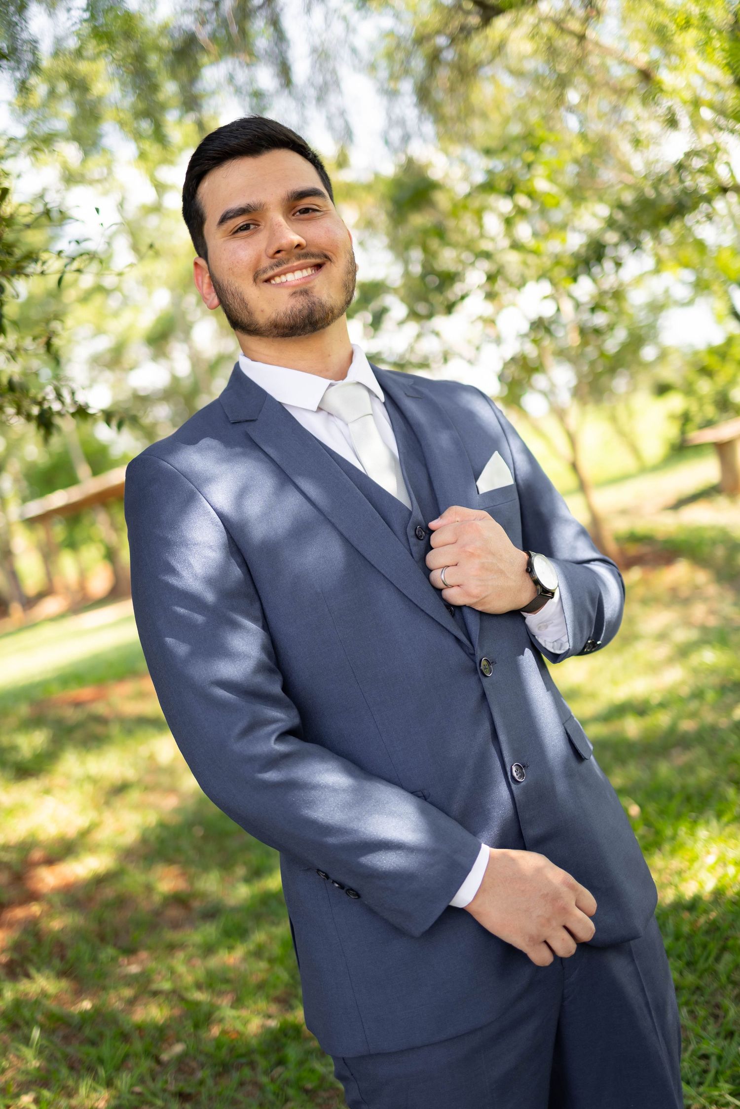 Smiling groom in a blue suit with white pocket square posing outdoors in a green park setting.