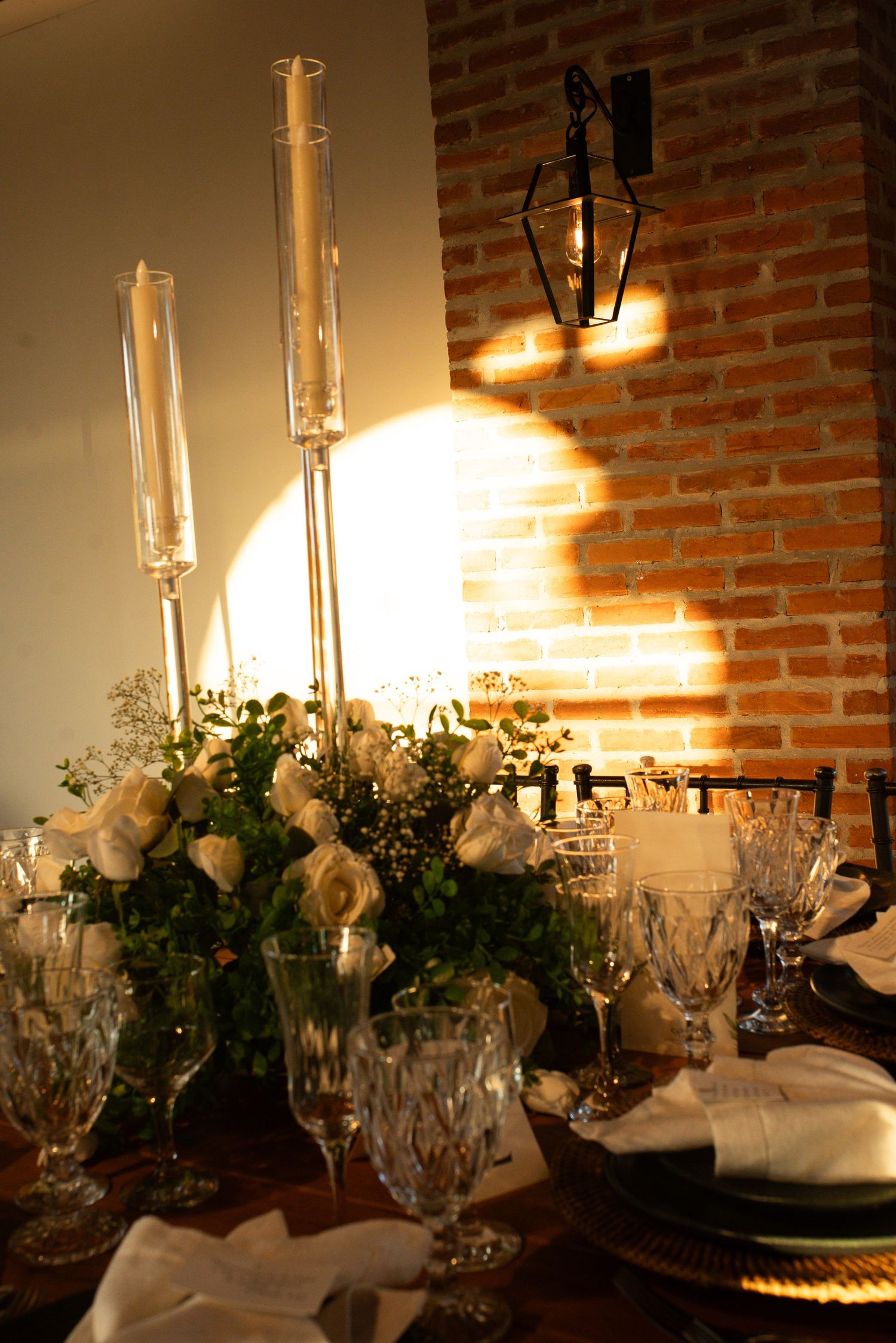 Elegant dining table with white roses, crystal glasses, candelabras, and warm wall sconce light against exposed brick wall.
