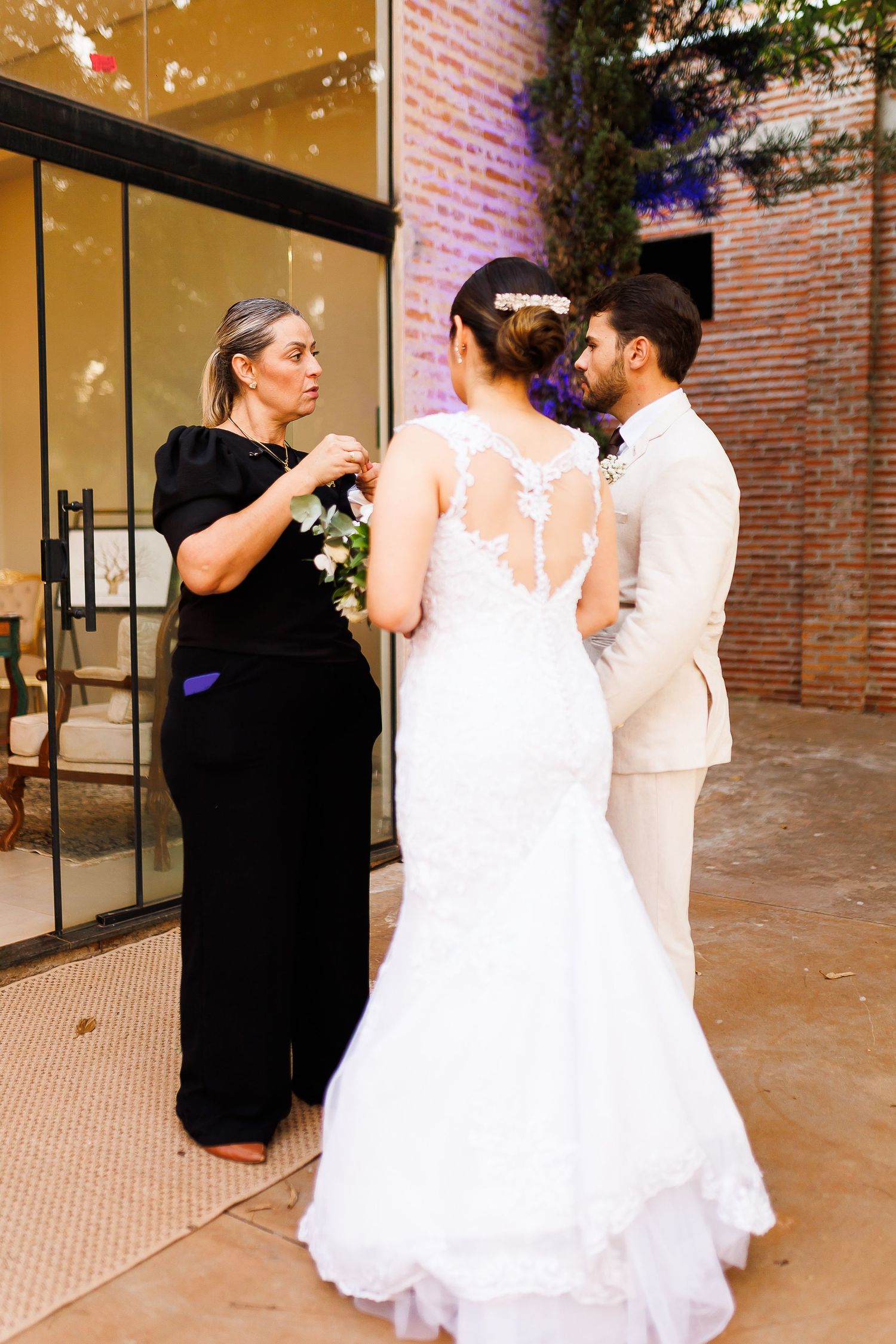 Wedding coordinator adjusts bride's bouquet as groom stands nearby at an outdoor venue.
