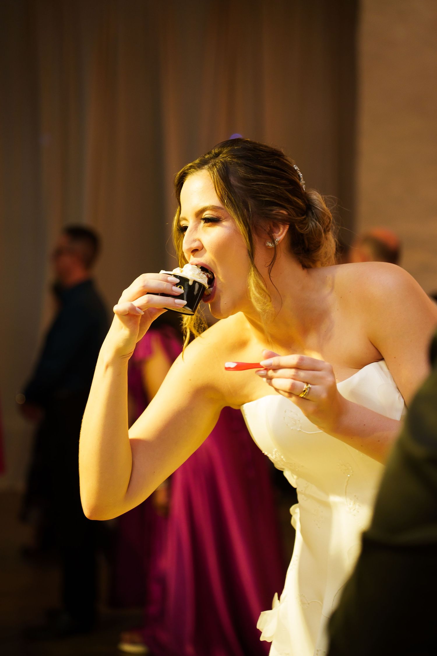 Bride in white strapless gown eating wedding cake at reception with guests in background.