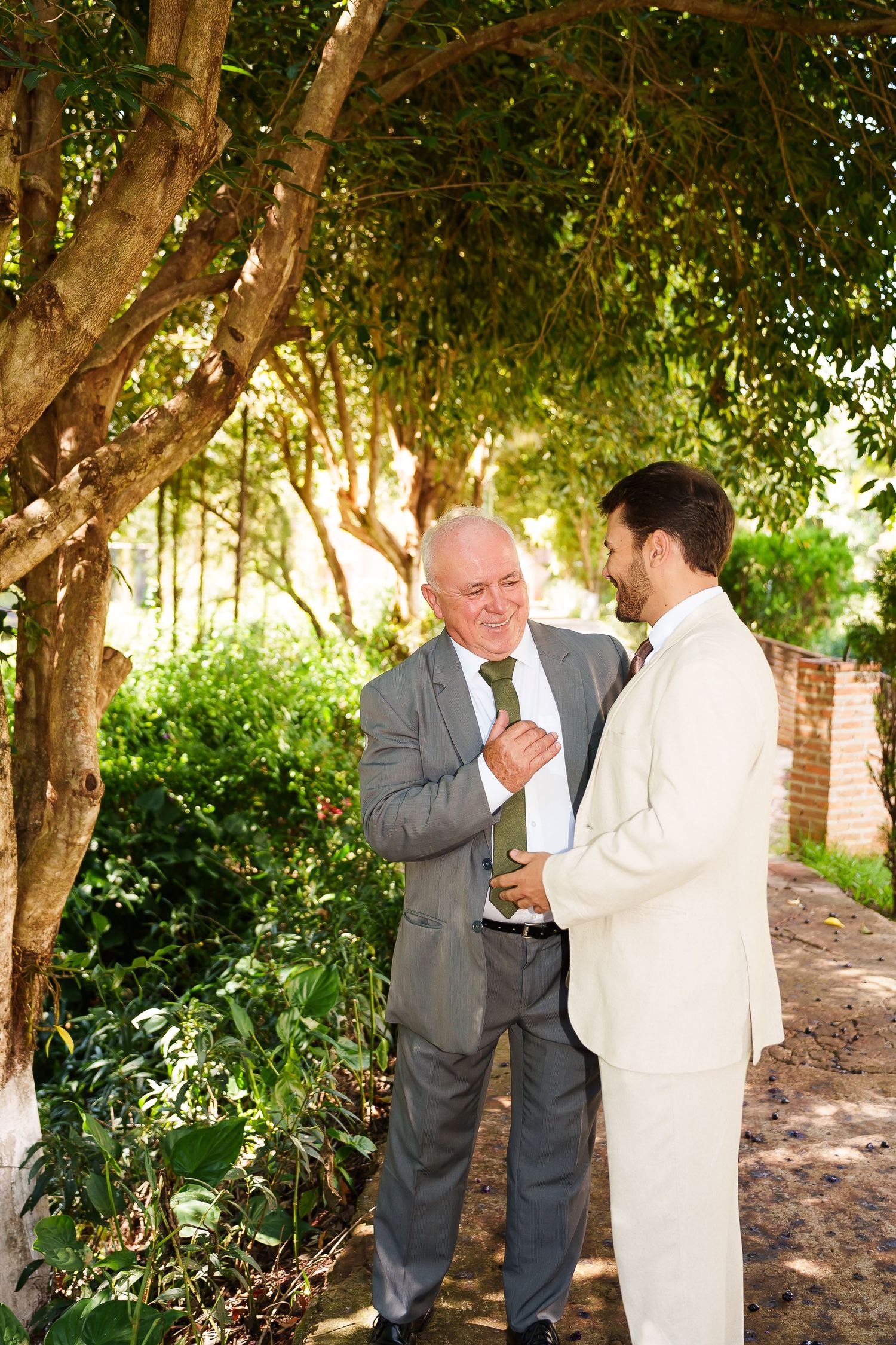 Two men in suits share a moment in a lush garden, with an older man adjusting the younger mans cufflink.