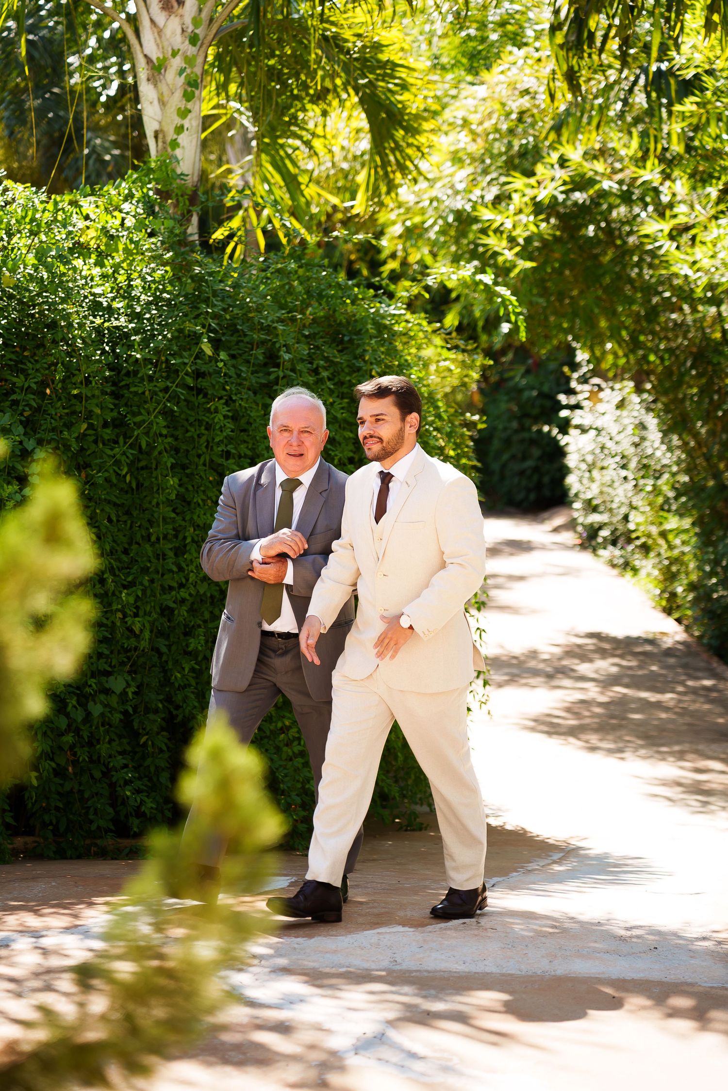 Two men in suits walk together on a garden path surrounded by lush green hedges and palm trees.