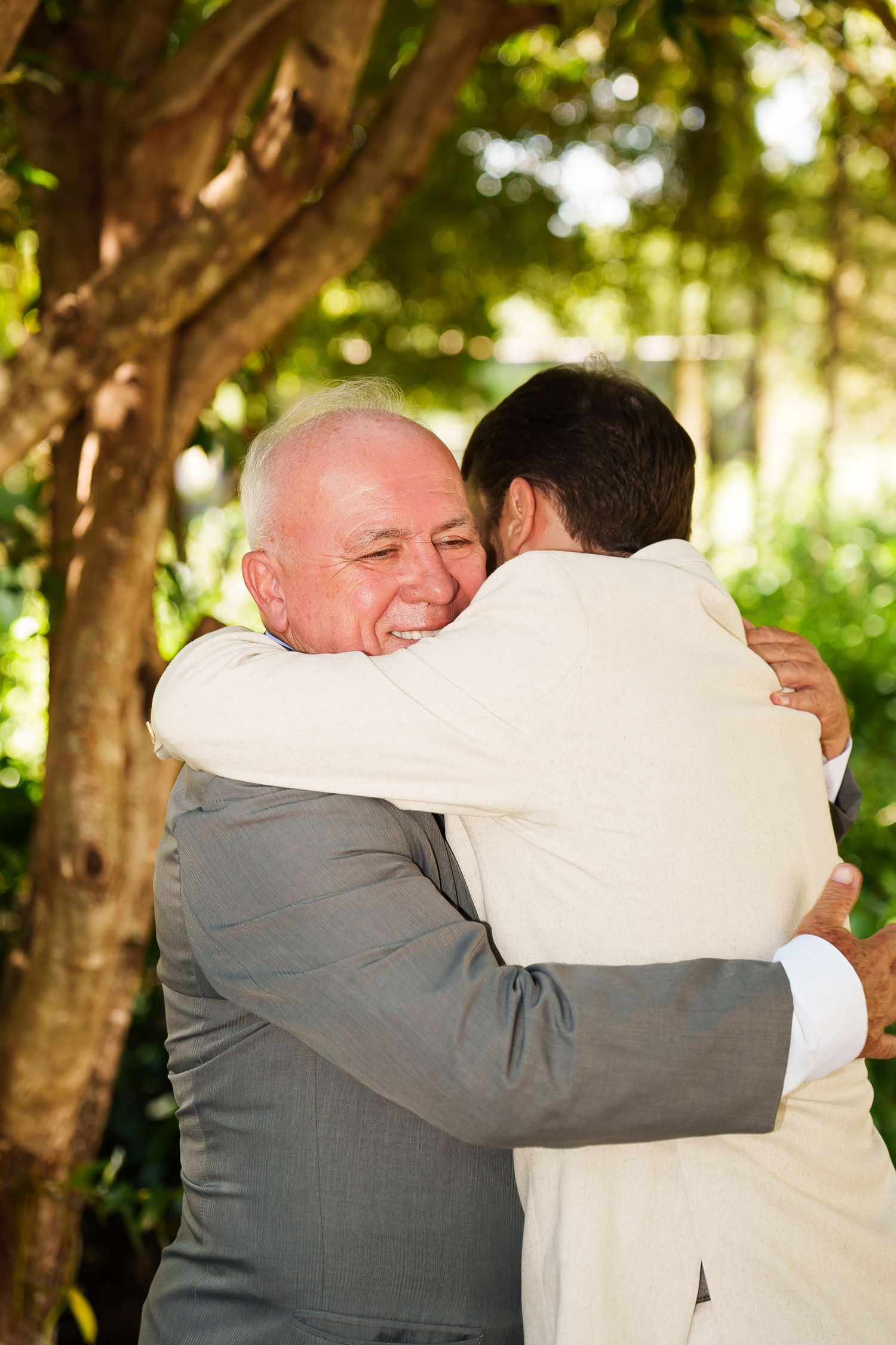 Two men in suits share a warm embrace outdoors at a wedding ceremony, surrounded by lush green trees.