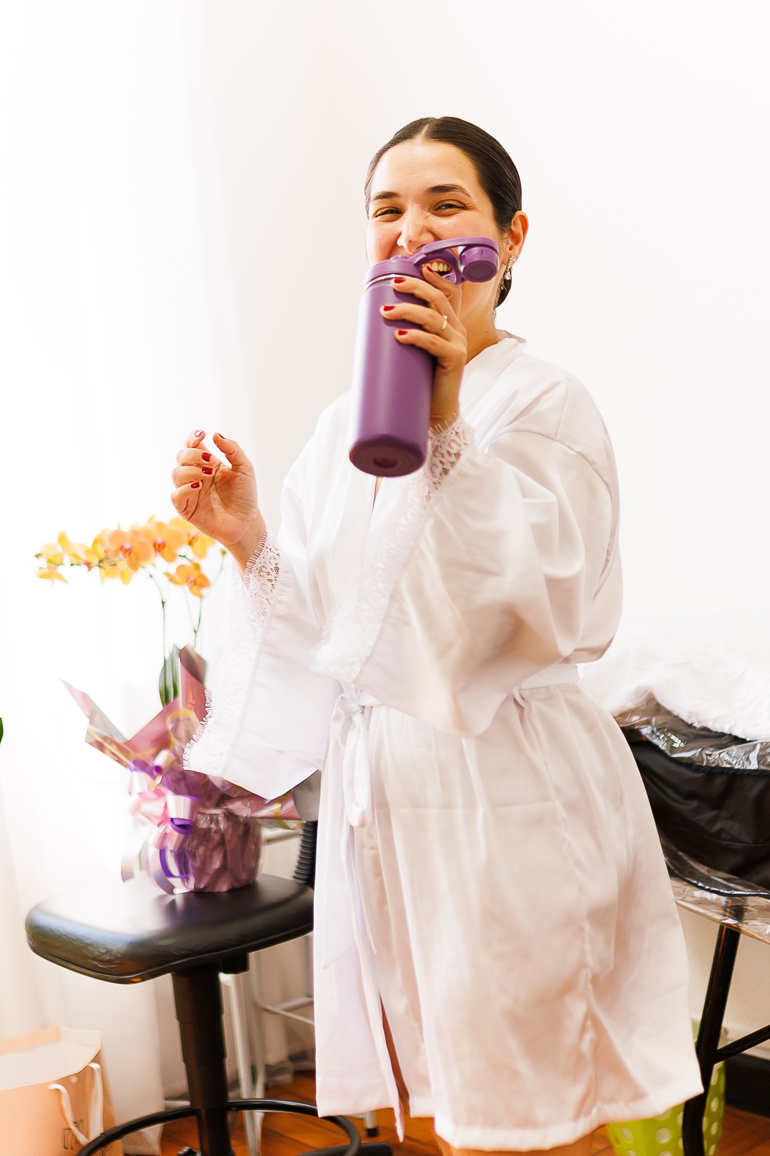 A woman in a white robe sips from a purple tumbler while holding hair curlers in a bright room.