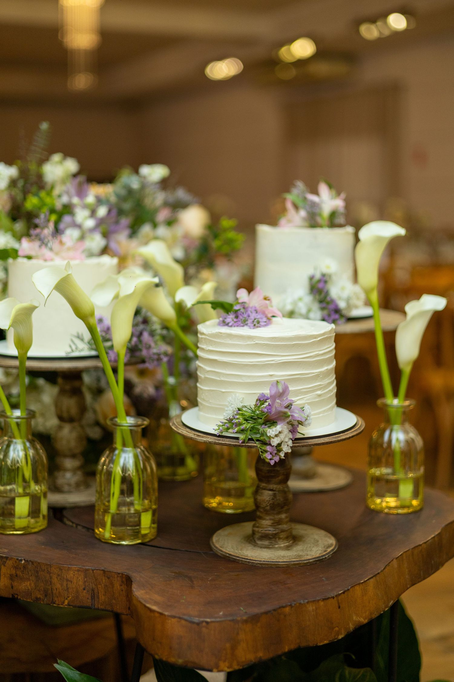 Elegant white wedding cakes on wooden table with purple flowers and green glass bud vases at a reception.