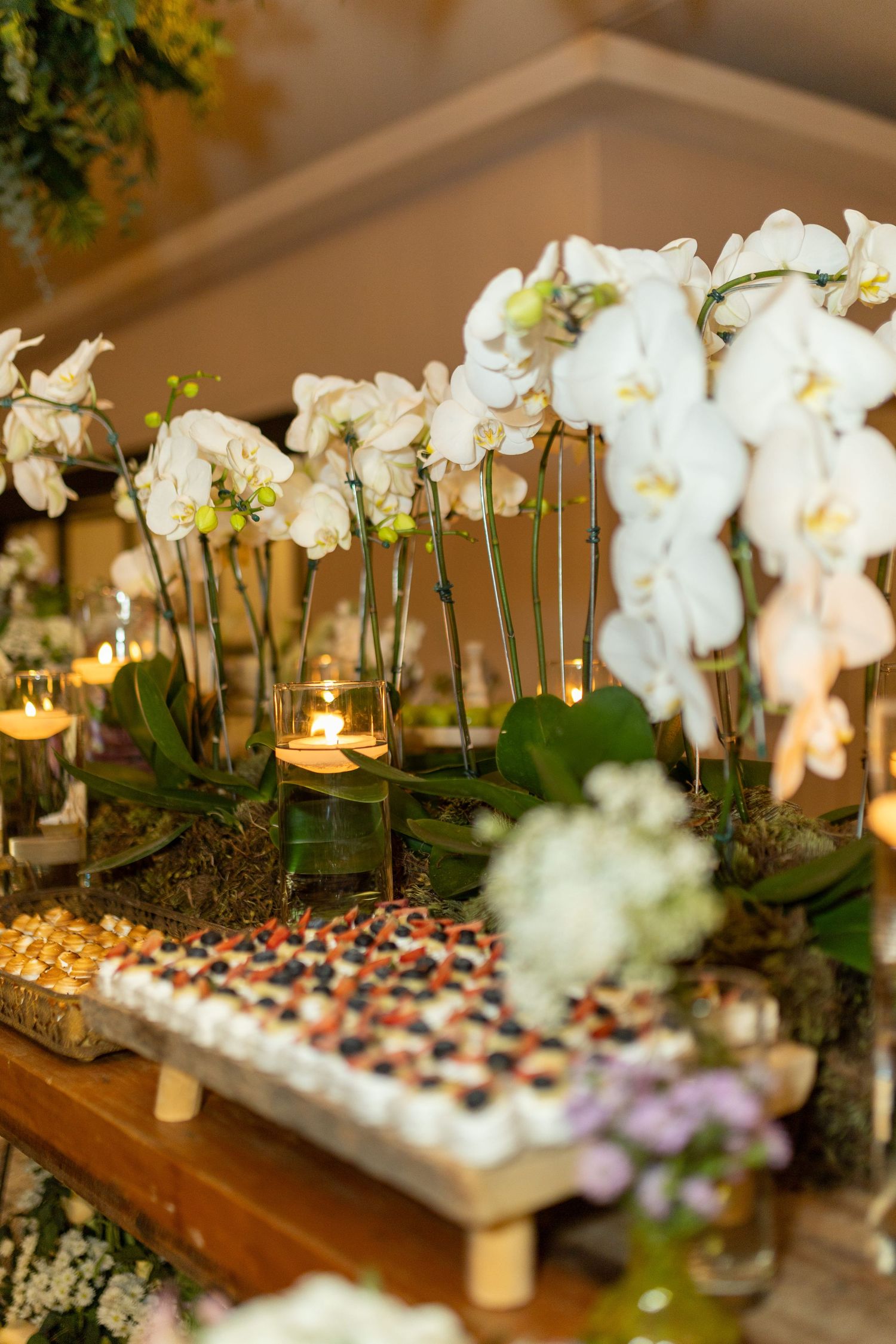 Elegant wedding dessert table with white orchids, candles, and assorted petit fours on a wooden display.