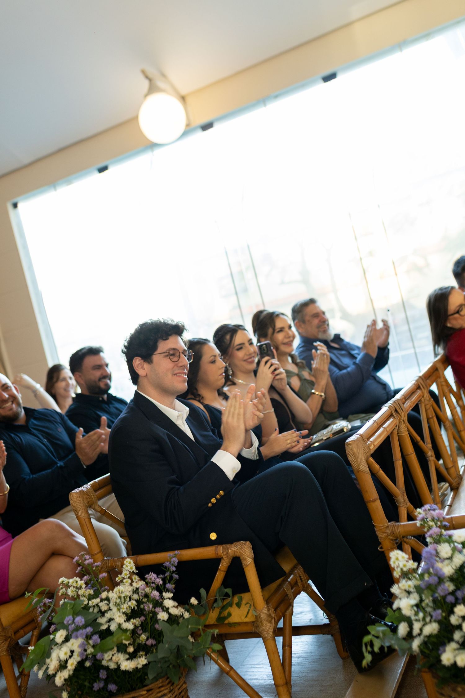 Wedding guests clapping and smiling during a ceremony, seated on bamboo chairs with floral arrangements nearby.