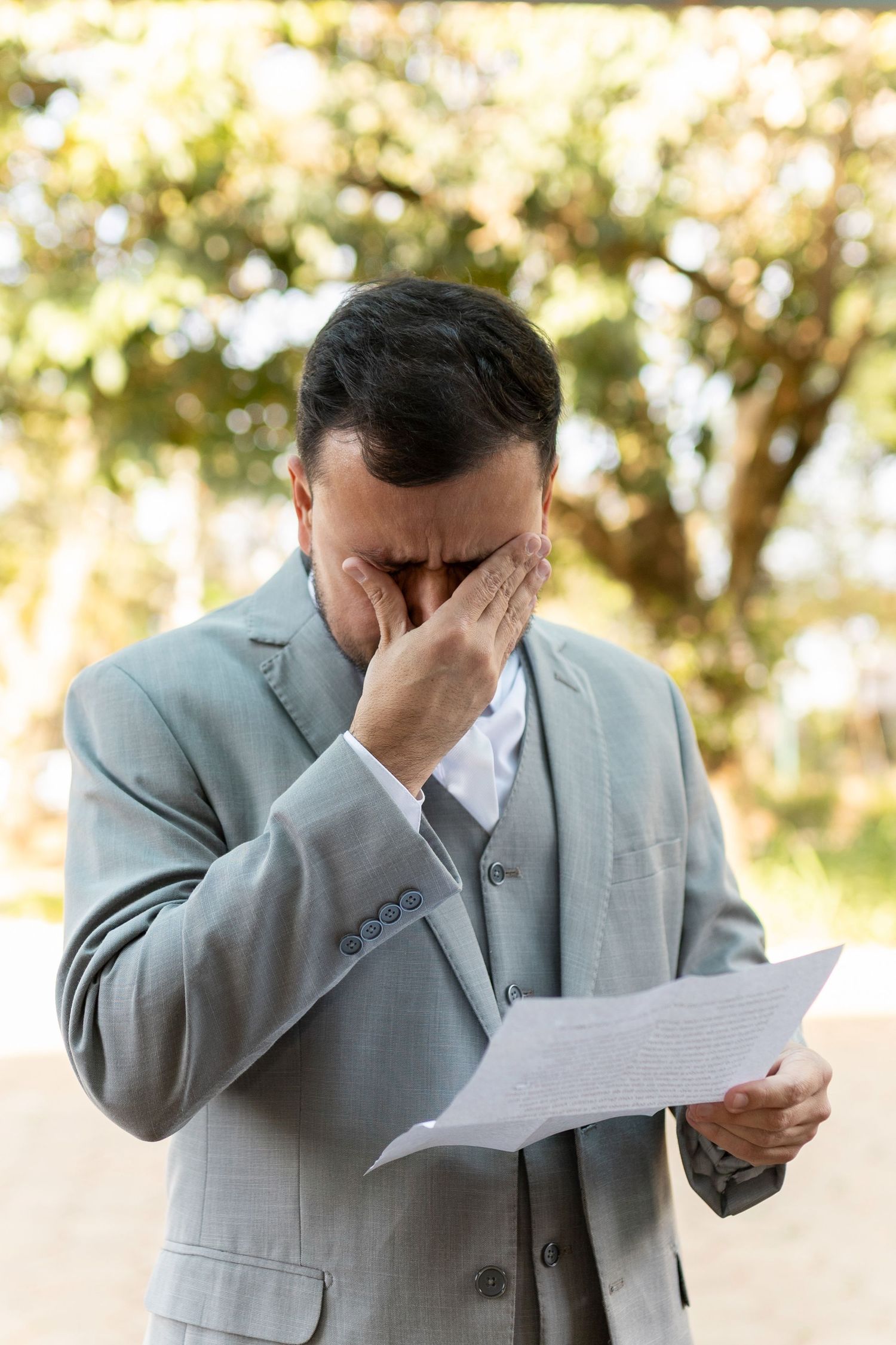 Stressed businessman in gray suit covering face while reading important document outdoors in natural light.
