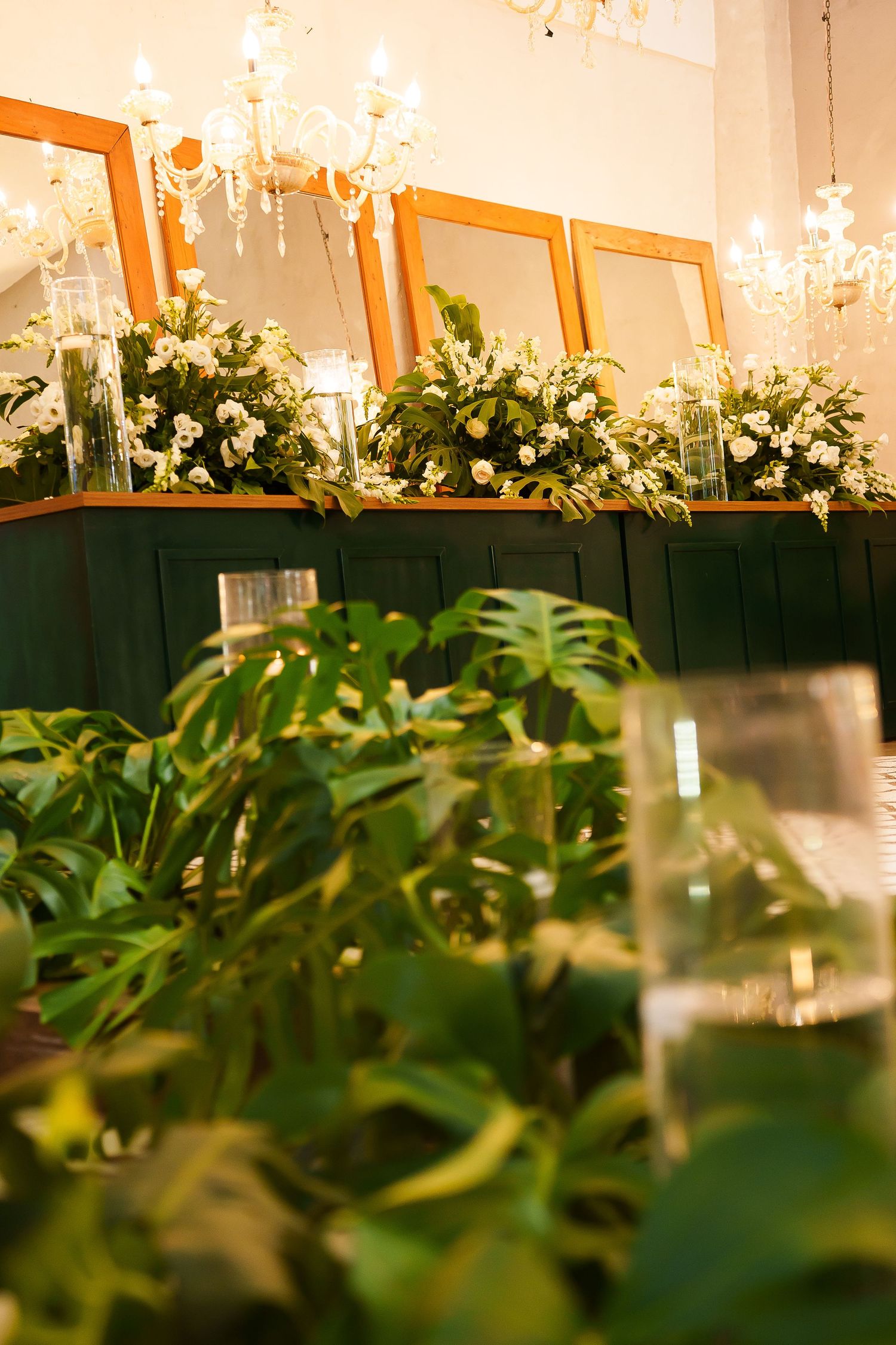 Elegant indoor space with lush tropical plants, white floral arrangements, gold-framed mirrors, and crystal chandeliers.