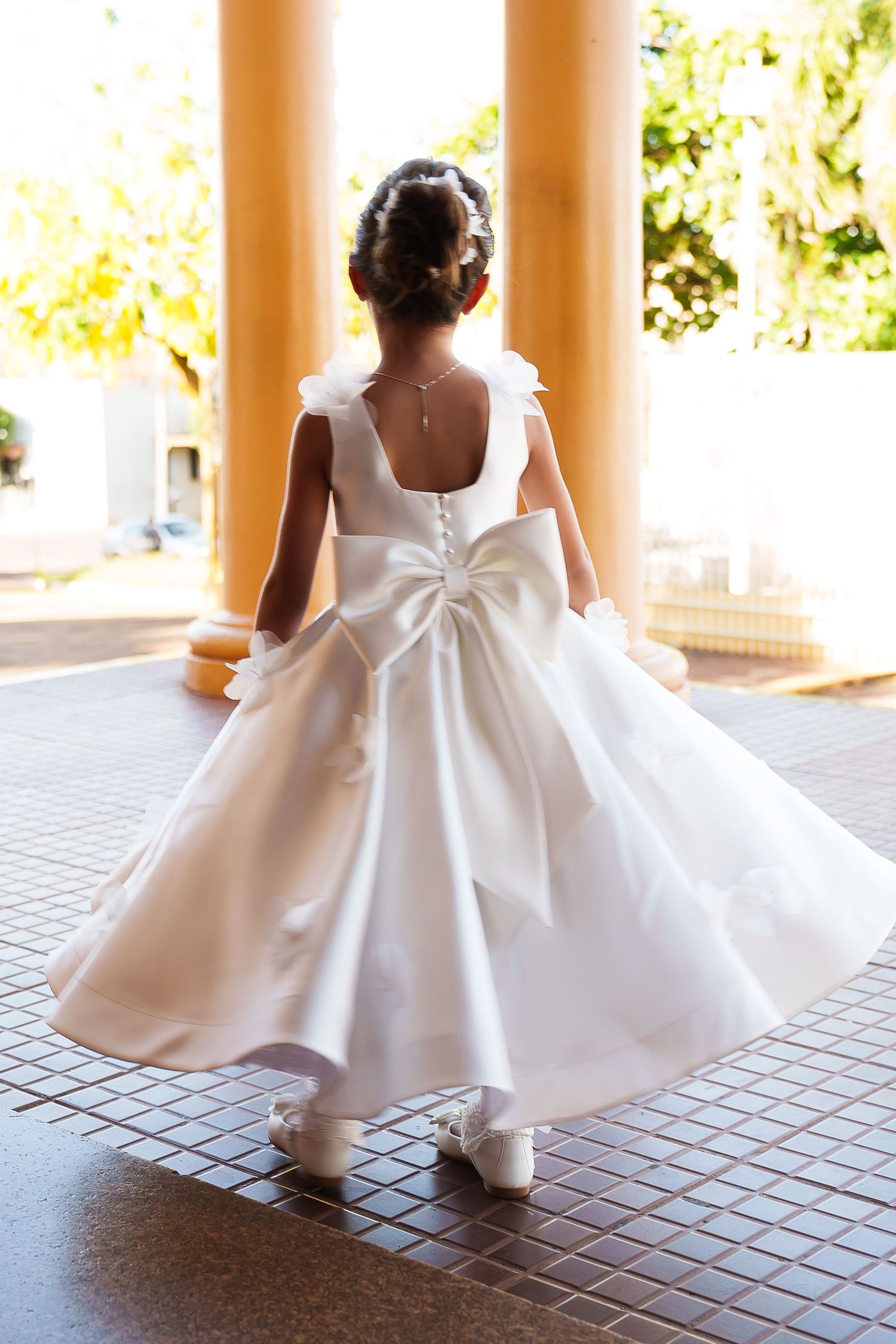 Back view of young girl in white satin flower girl dress with large bow and white shoes.