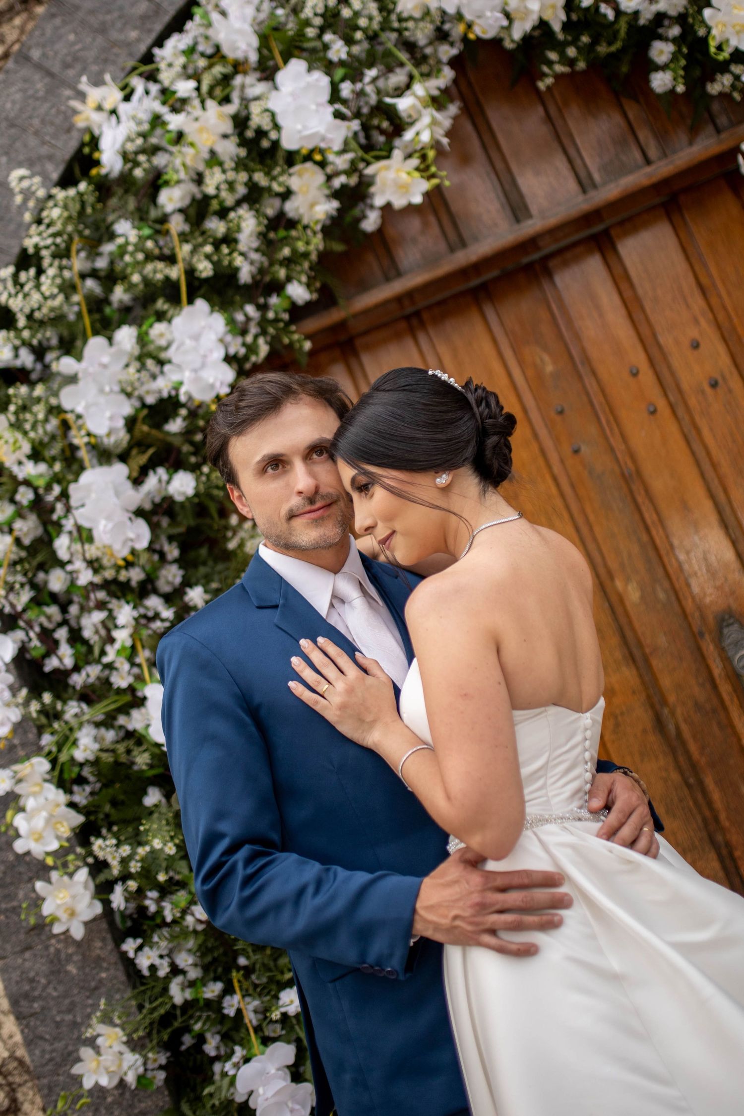 Bride and groom embracing before wooden doors adorned with white floral arch on their wedding day.