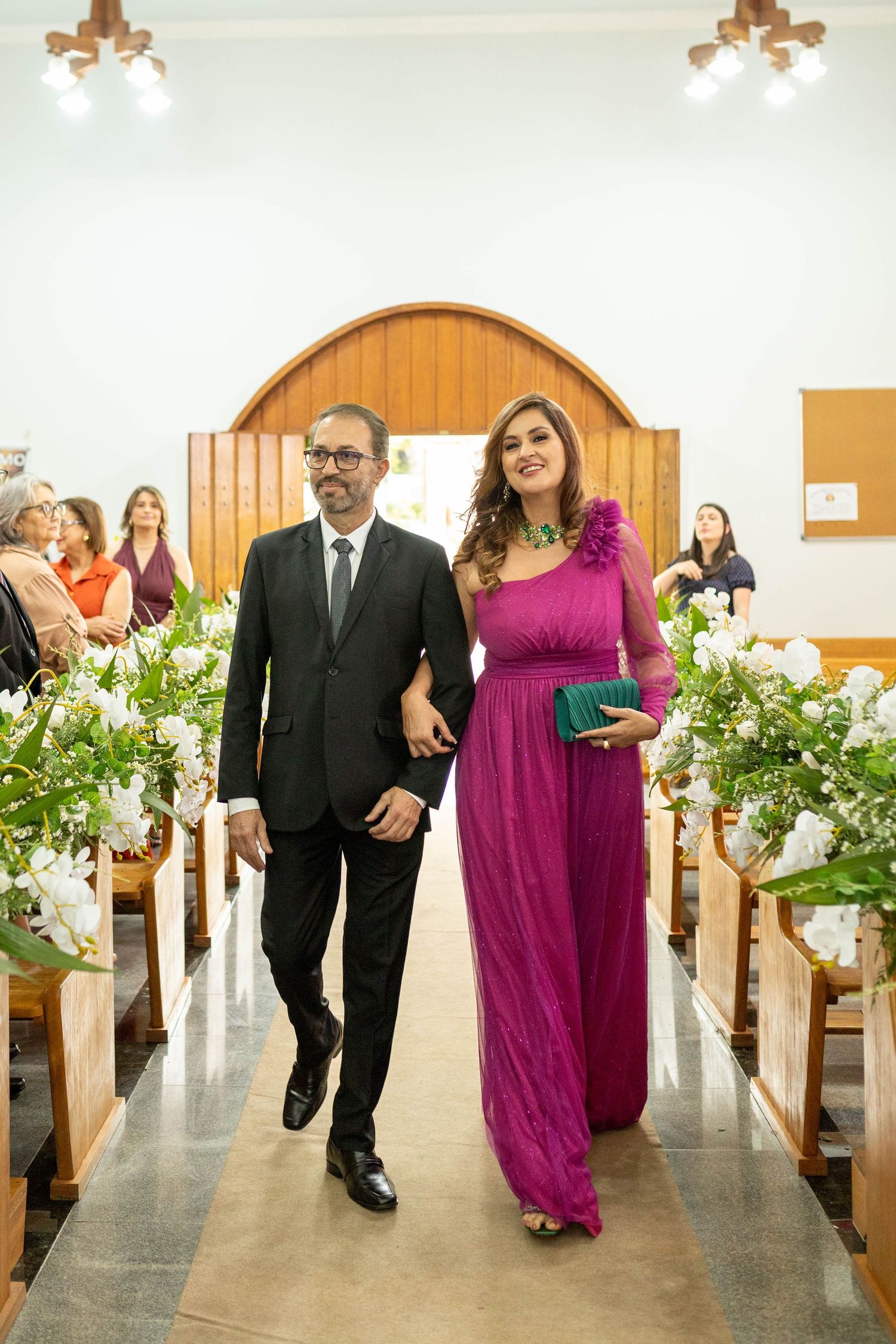 Couple walking down church aisle, man in black suit, woman in magenta gown holding green clutch, floral arrangements.