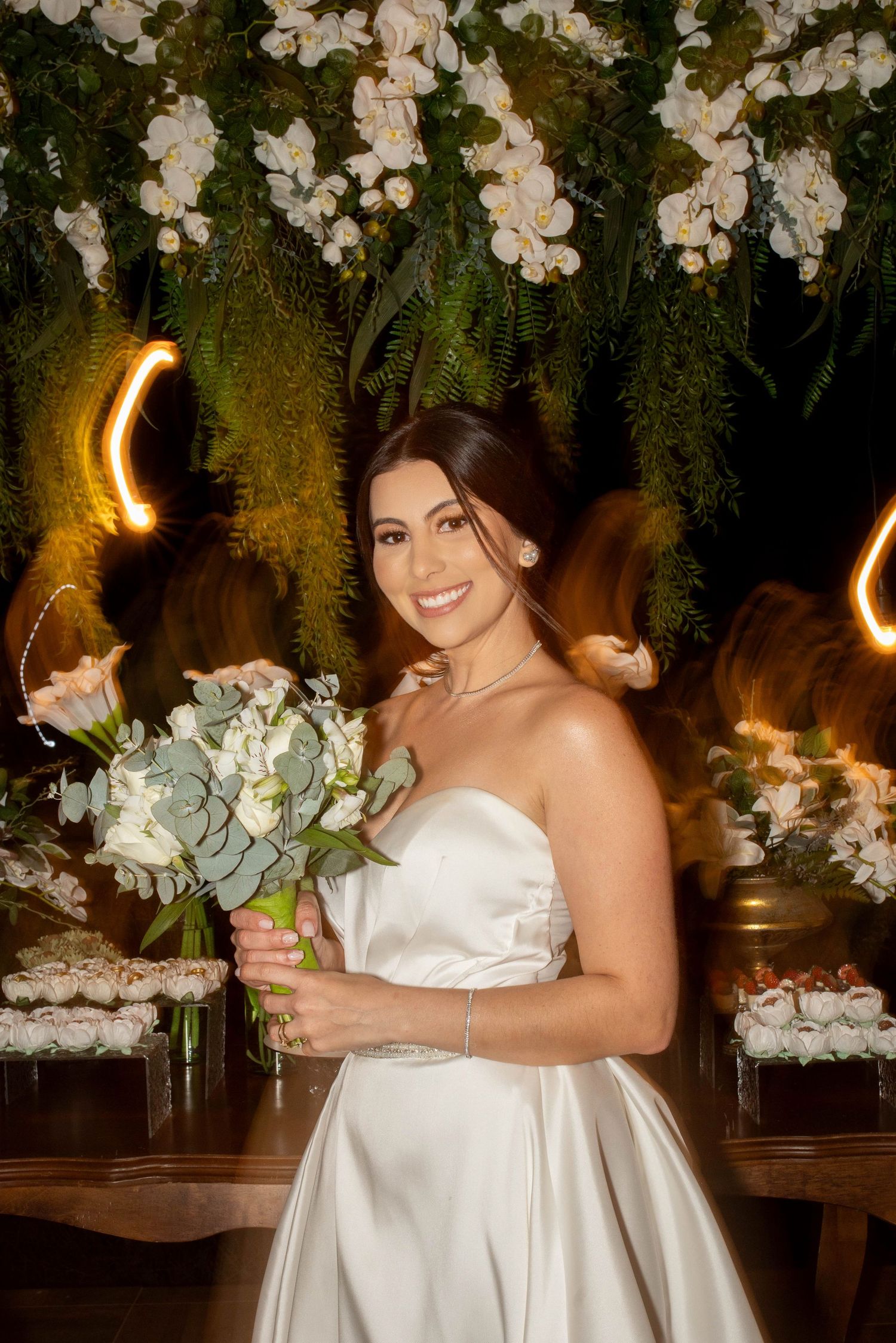 Smiling bride in strapless white satin gown holding white floral bouquet, posing before lush greenery and floral backdrop.