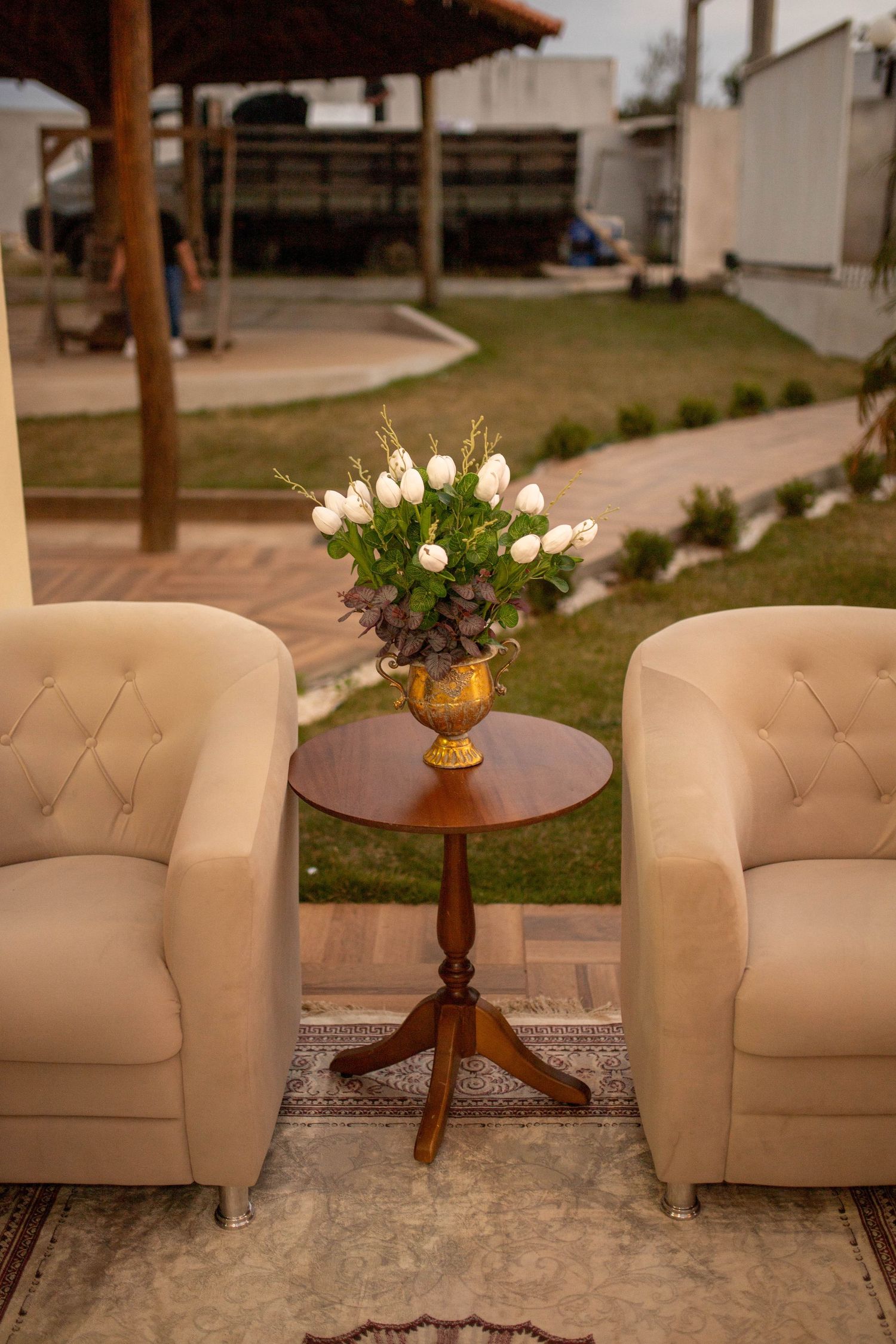 Two beige tufted armchairs flank a wooden pedestal table with white tulips in a gold vase outdoors.