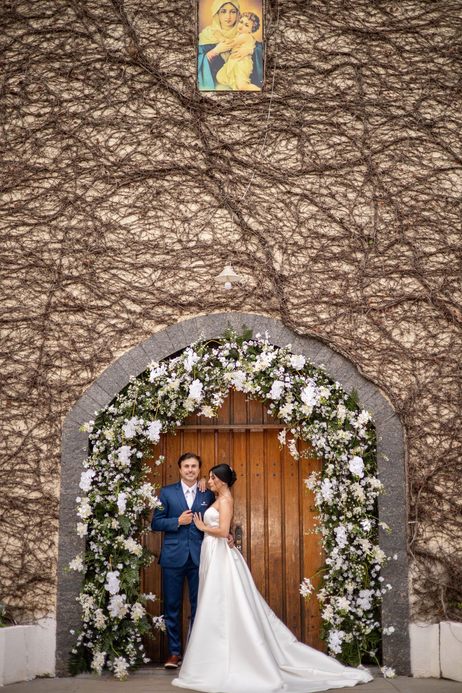 Bride and groom stand under a floral arch adorned with white flowers at a church entrance with a religious icon above.