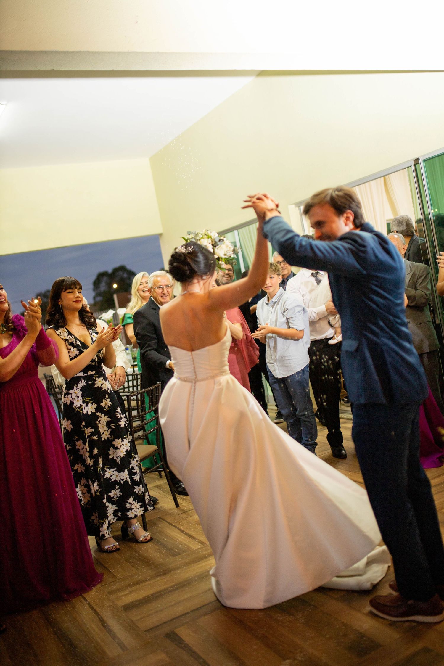 Bride and groom share their first dance at wedding reception while guests applaud and watch in formal attire.