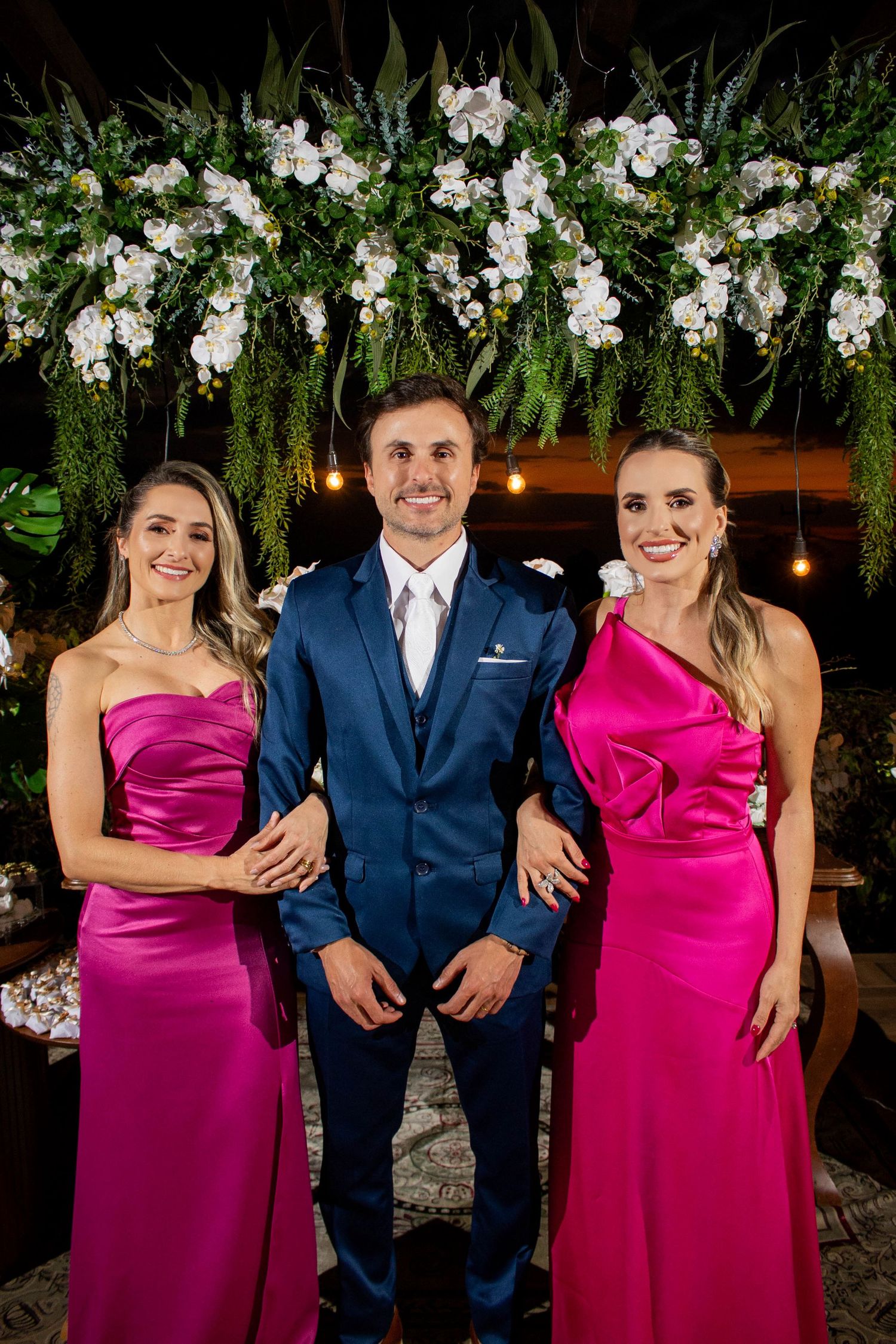 Man in blue suit poses with two women in pink satin gowns under floral arch at elegant wedding celebration.