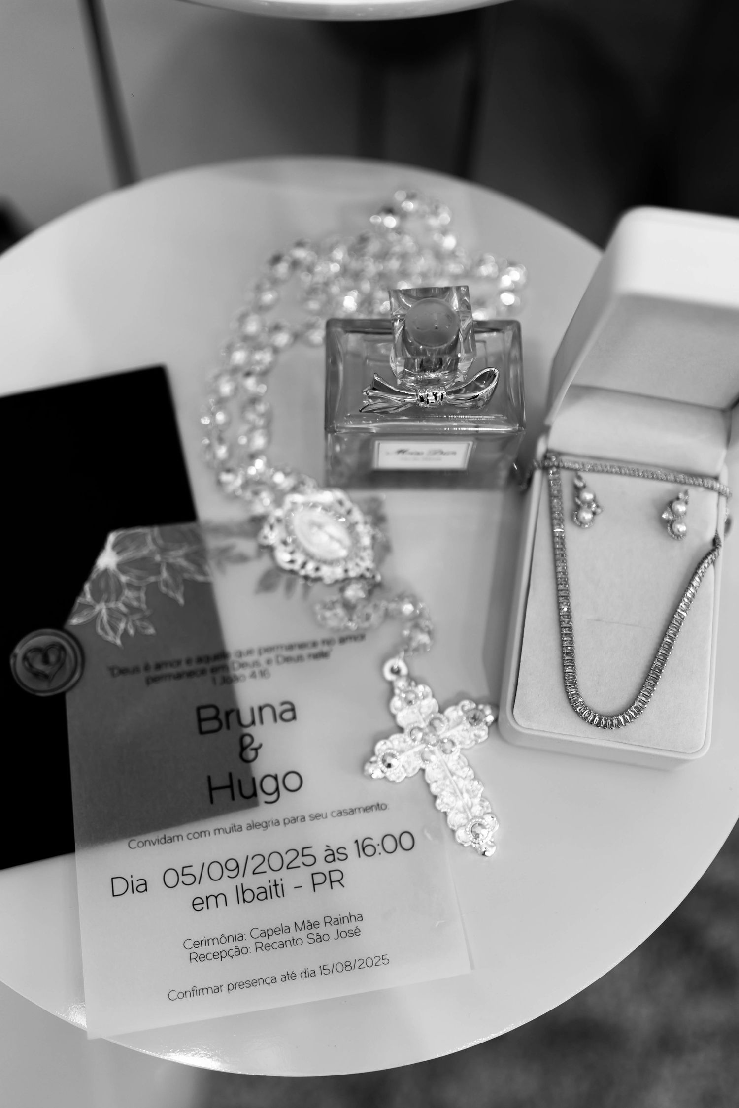 Black and white photo of Bruna and Hugo wedding invitation with rosary, perfume bottle, and diamond jewelry on white table.