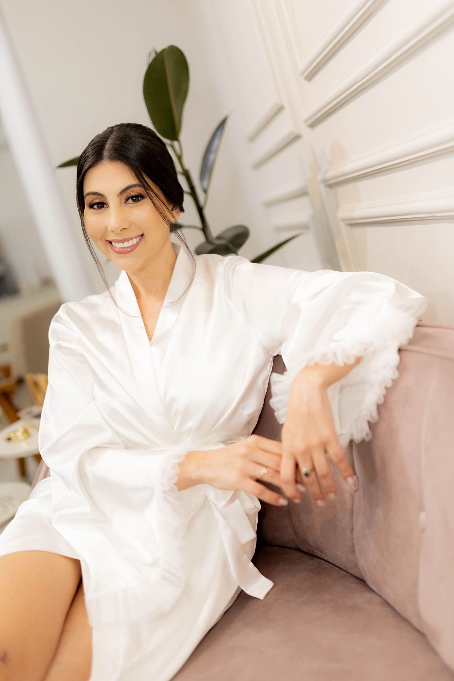 A smiling brunette woman in a white satin robe sits elegantly on a pink velvet sofa in a bright, modern room.