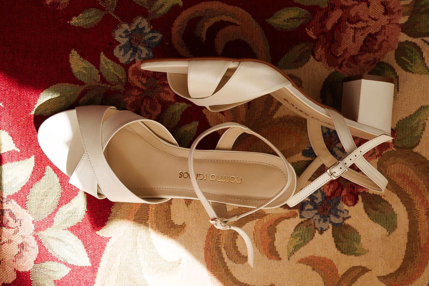 Elegant white strappy block heel sandals displayed on a floral red patterned rug.