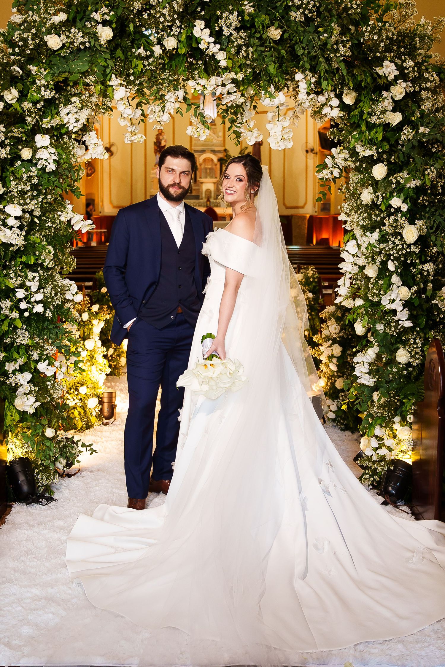 Bride and groom pose under floral arch with white flowers and greenery in elegant church wedding ceremony.
