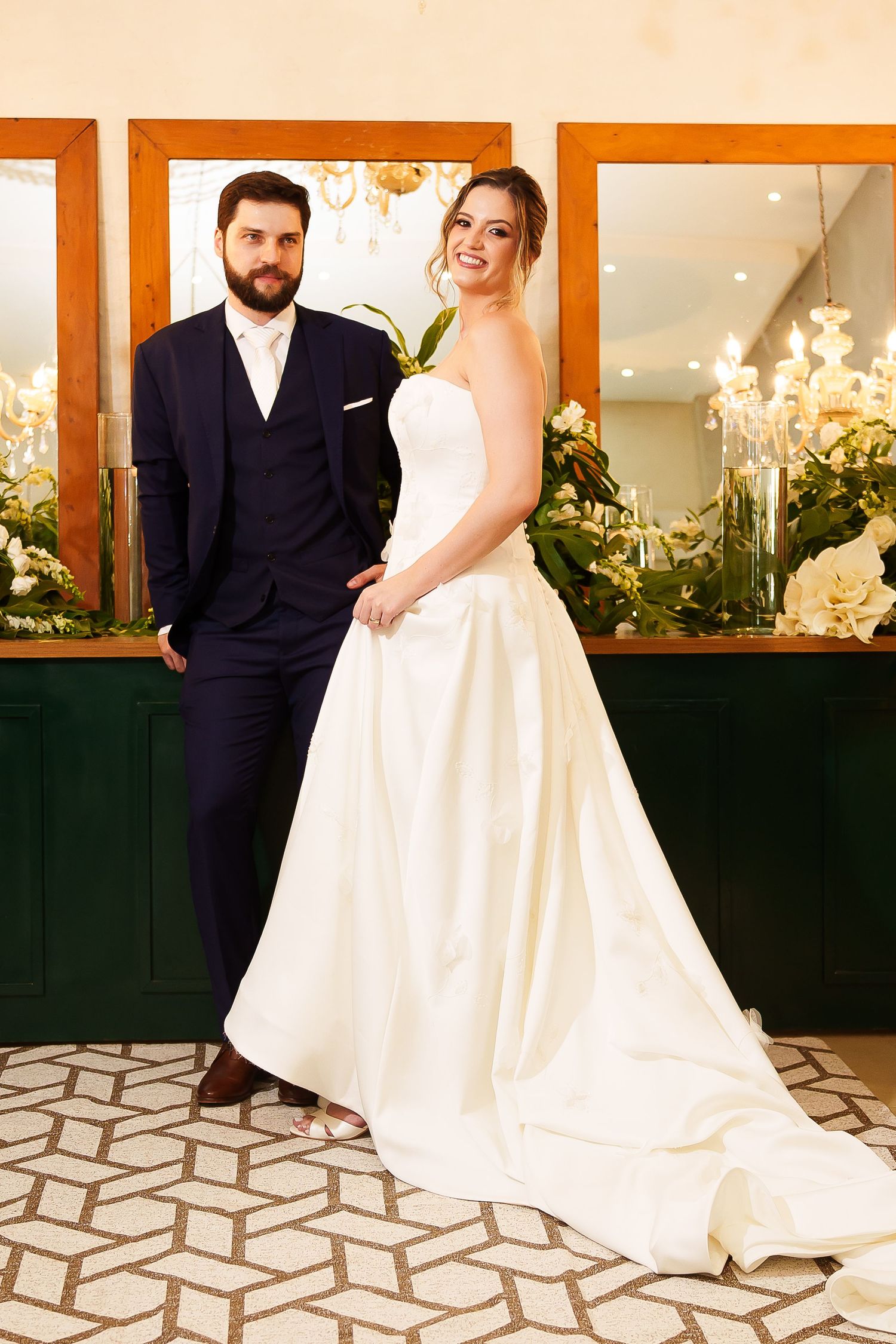 Elegant bride in white strapless gown and bearded groom in navy suit posing together in a decorated wedding venue.