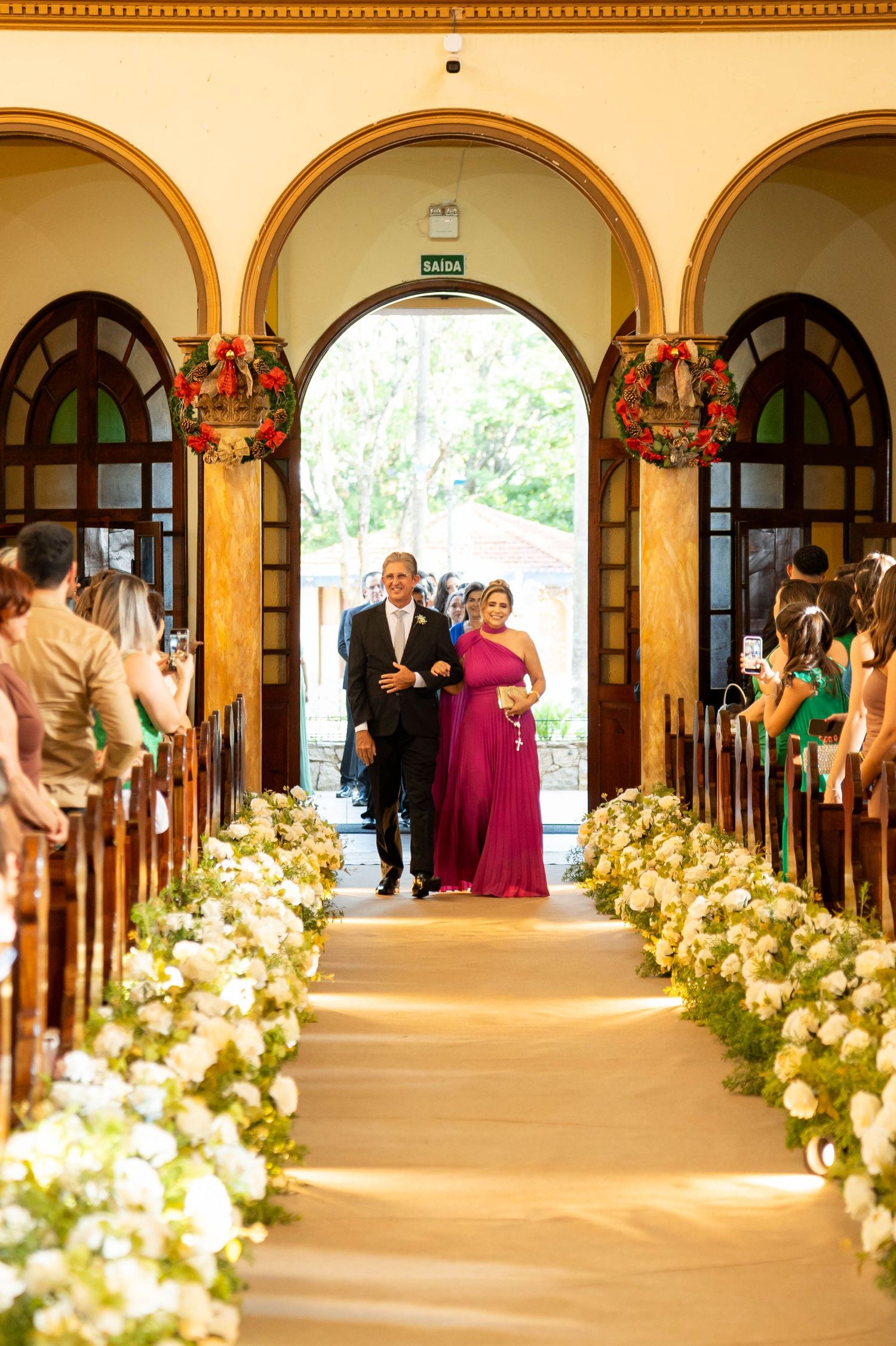 Elegant wedding ceremony aisle lined with white flowers, couple walking toward altar in a classic chapel with arched doorways.