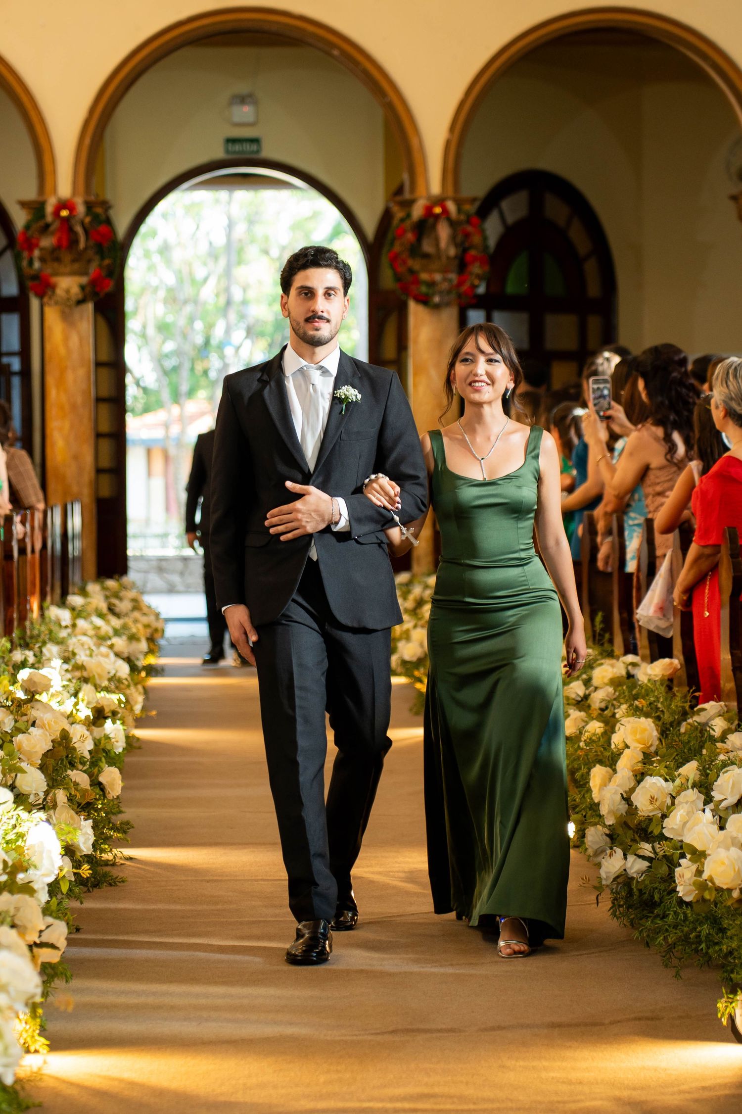 Elegant couple walking down a floral-lined wedding aisle, man in black suit, woman in green satin gown.