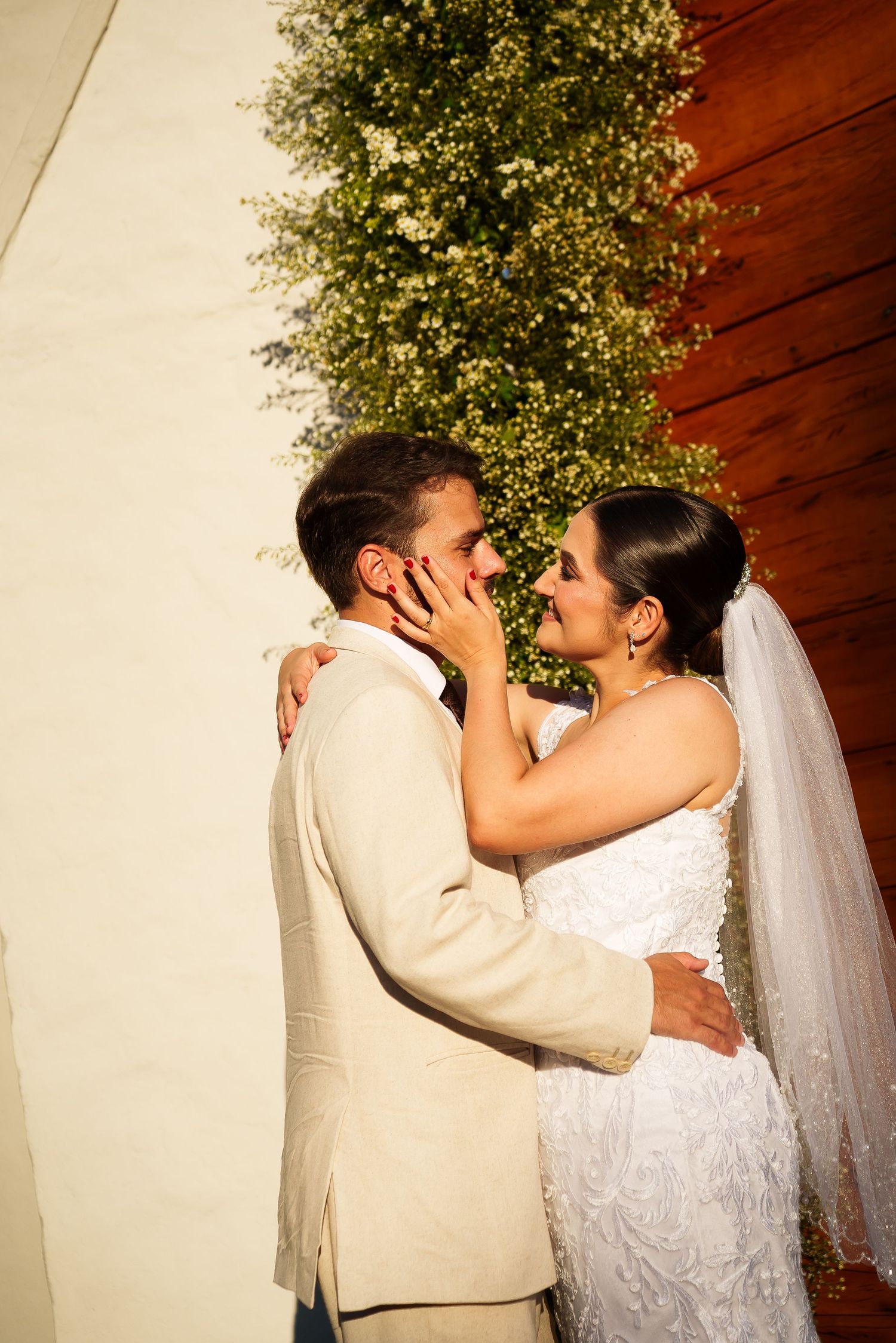 Bride and groom share a romantic moment, with bride in white lace gown and veil touching grooms face outdoors.