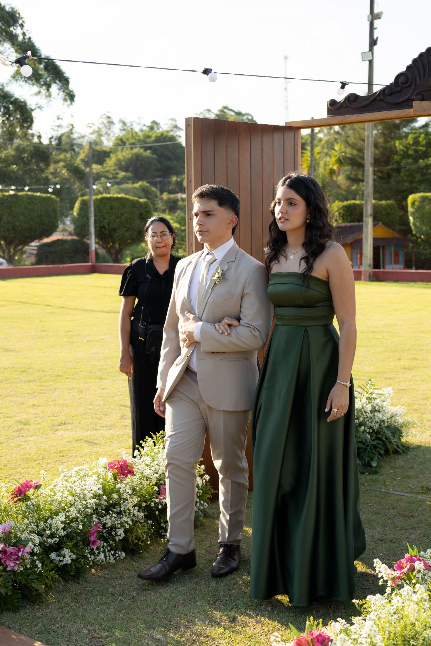 Elegant outdoor wedding ceremony with couple in beige suit and green gown standing on lush lawn with floral decorations.