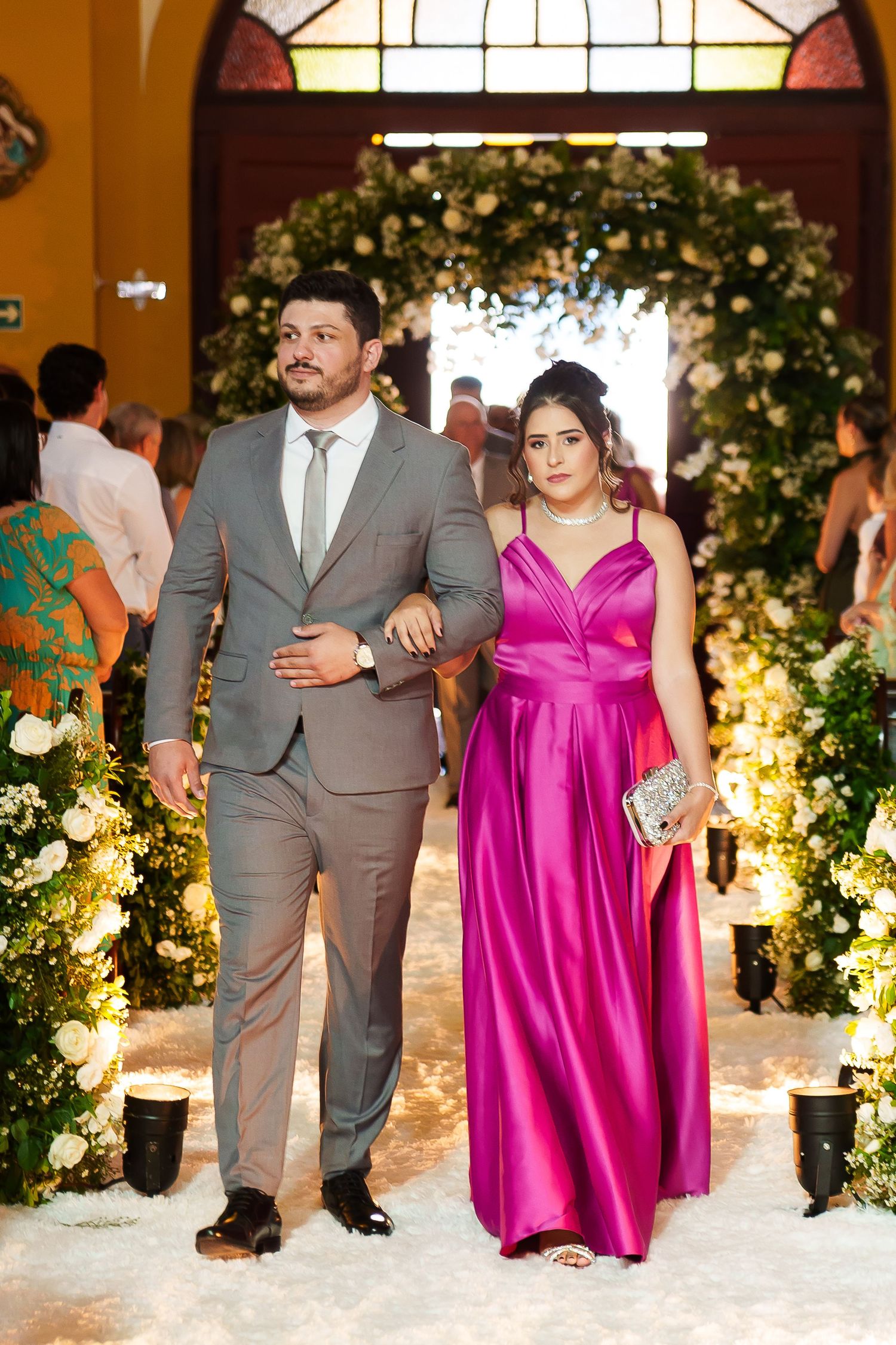 Elegant couple posing at a wedding venue, man in gray suit and woman in pink satin gown, surrounded by floral decor.
