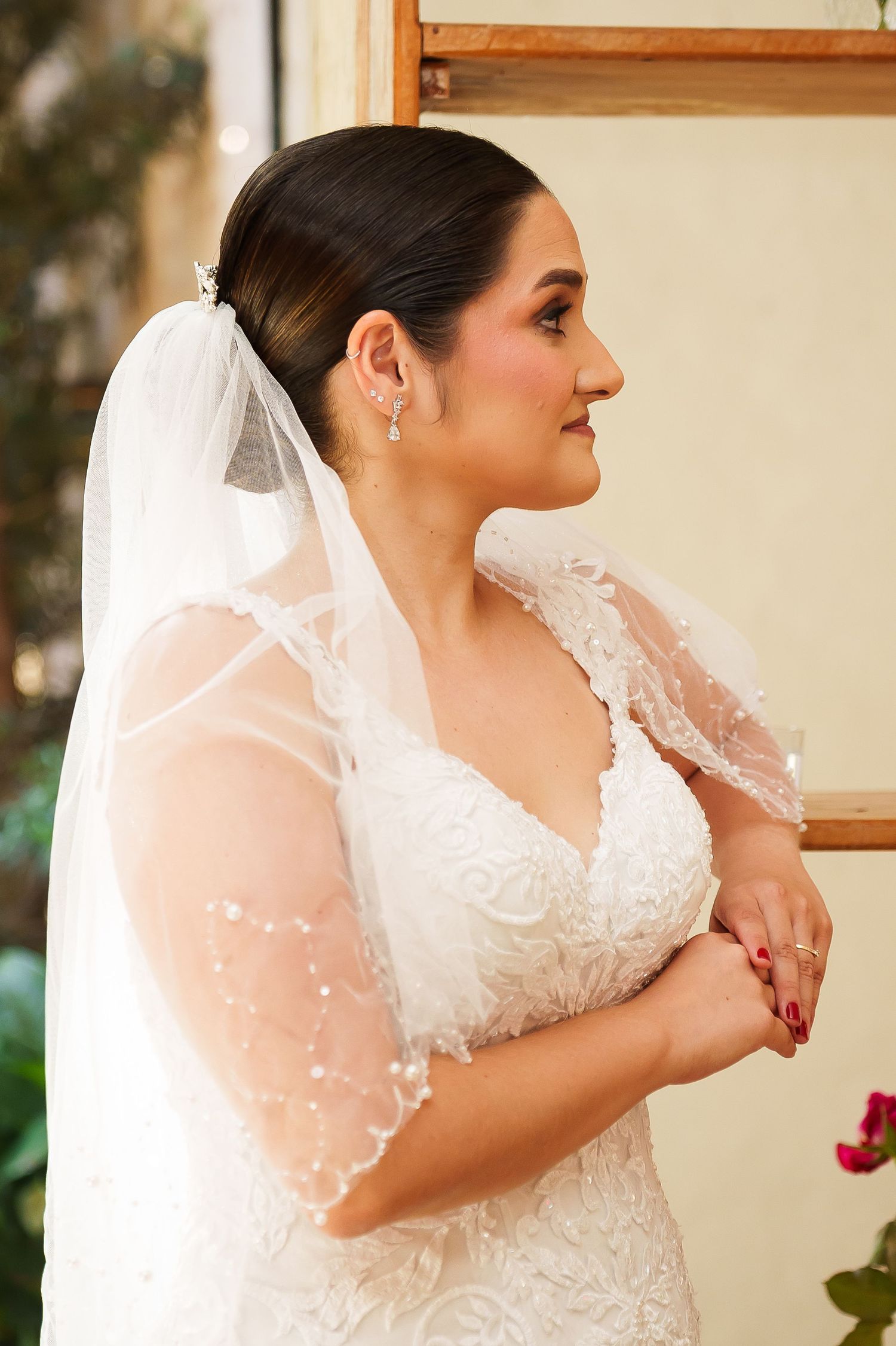 Bride in lace wedding dress and pearl-trimmed veil with elegant updo, wearing drop earrings, smiling in profile view.