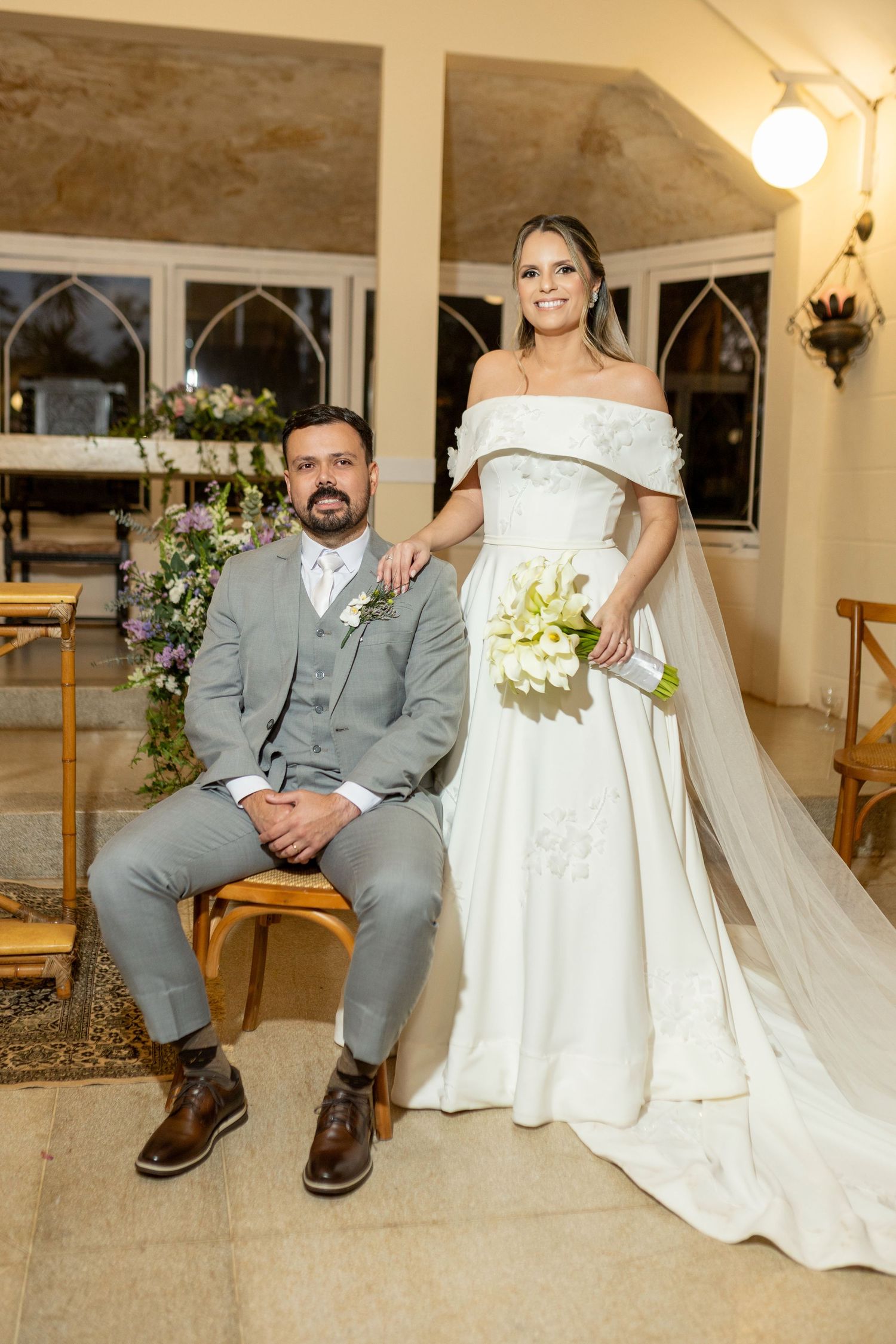 Bride in off-shoulder white gown holding green bouquet stands beside groom in gray suit seated in church.