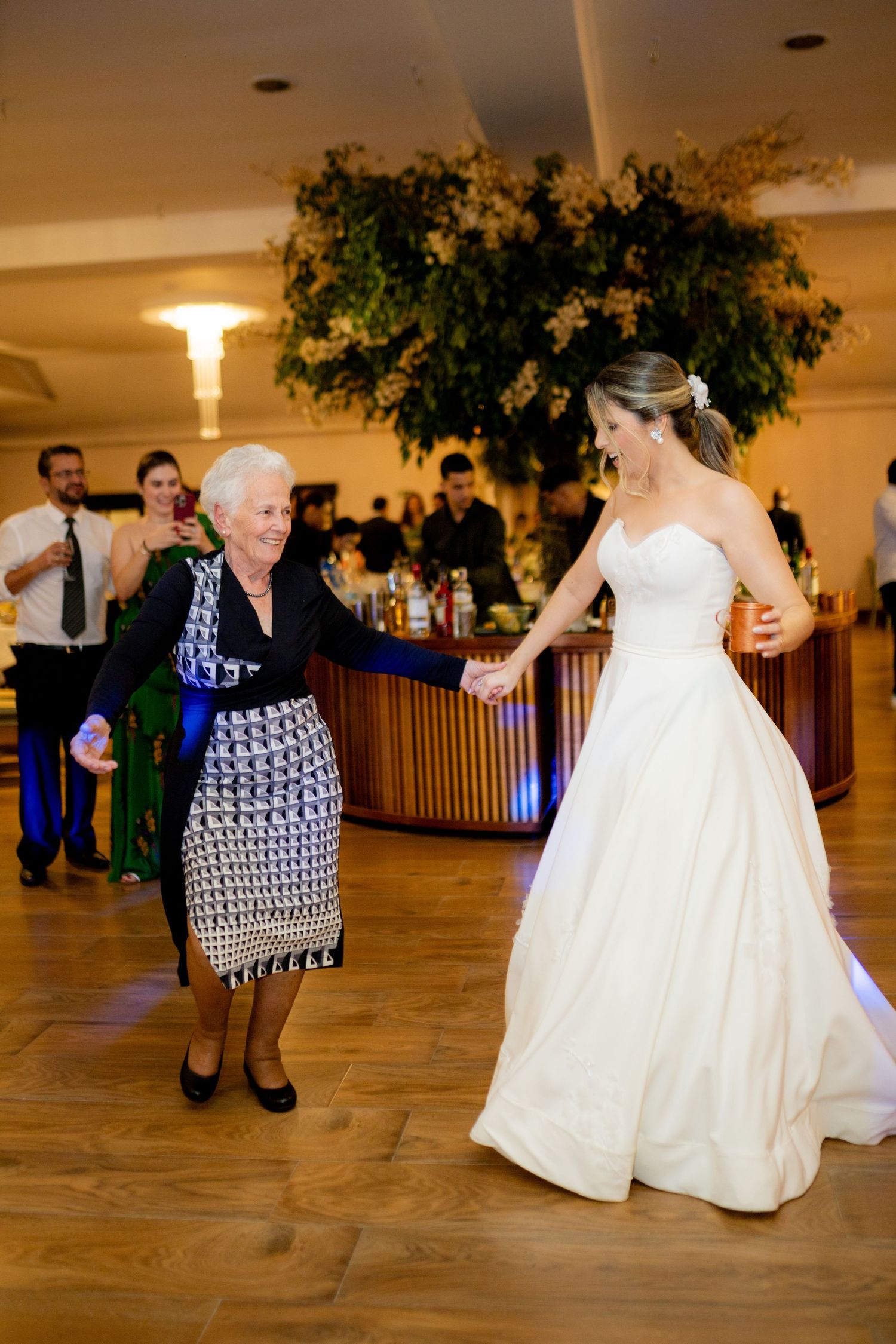 Bride in white gown dancing joyfully with elderly woman in patterned dress at wedding reception.