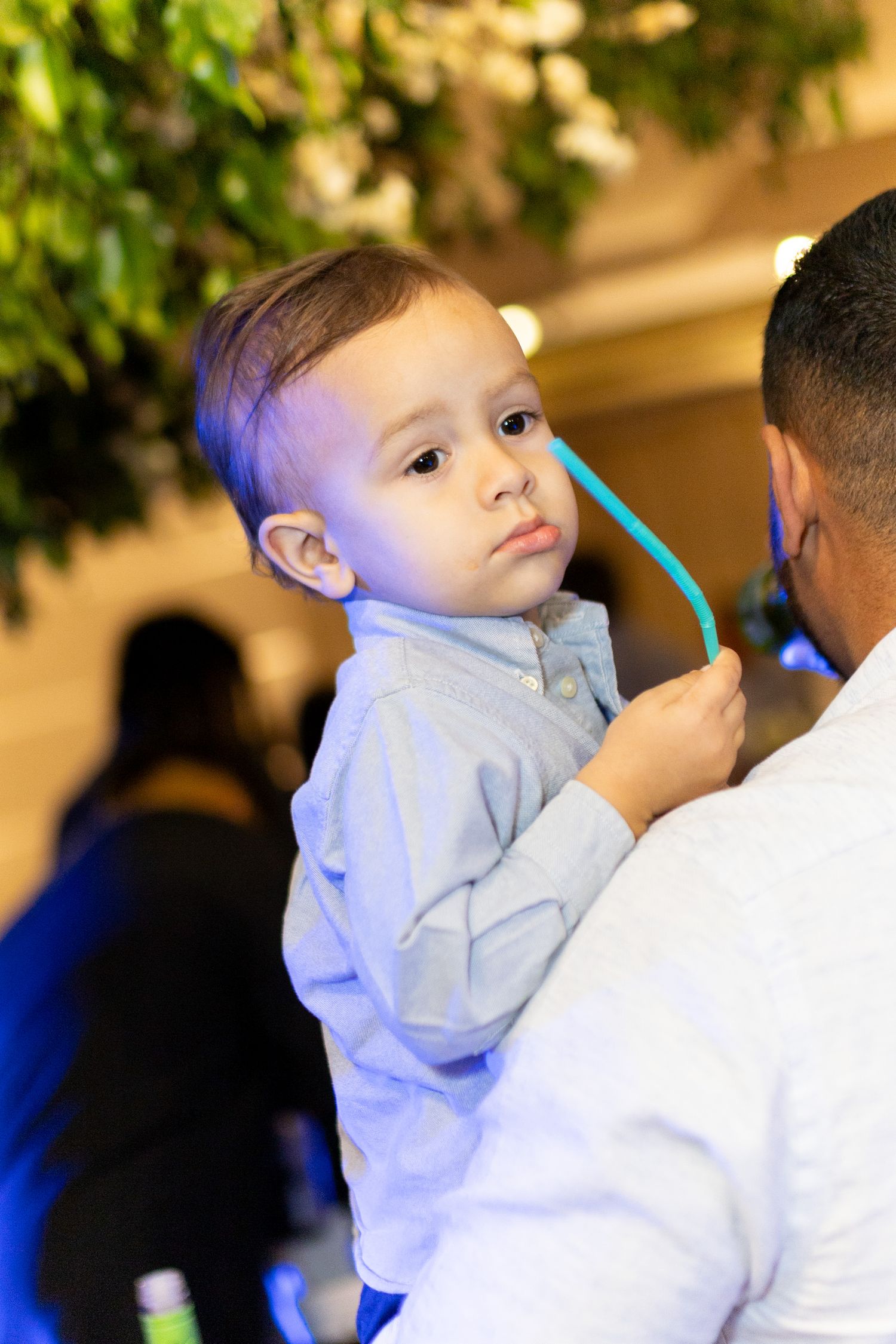 A toddler boy in a blue shirt is held by an adult while playing with a teal straw at an indoor event.